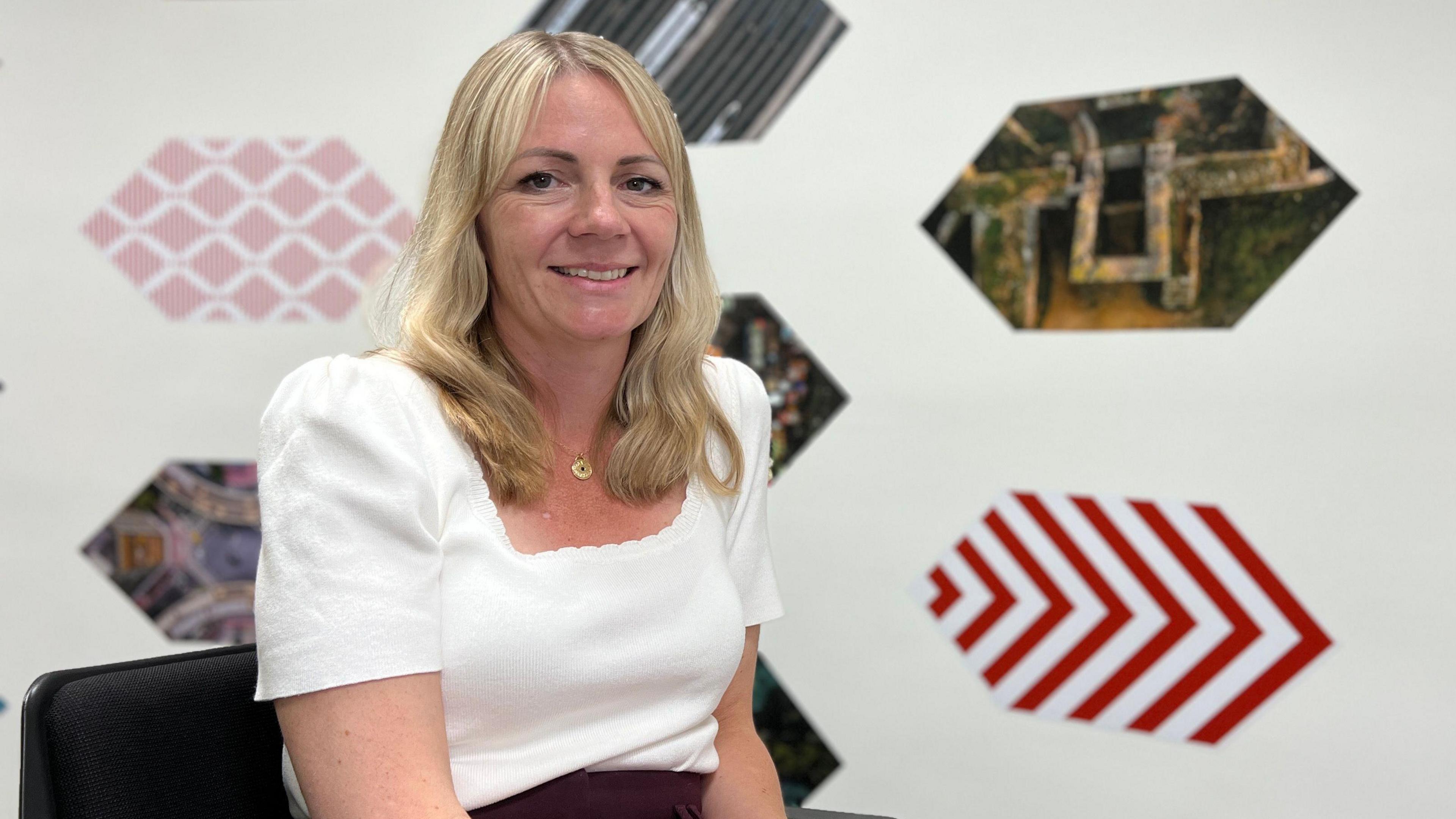 Chloe is seated on a black chair, wearing a white top and maroon skirt, hands resting on their lap. The background features hexagonal images of landscapes, cityscapes, and red and white chevron patterns. The word 'Open' is partially visible on the right.