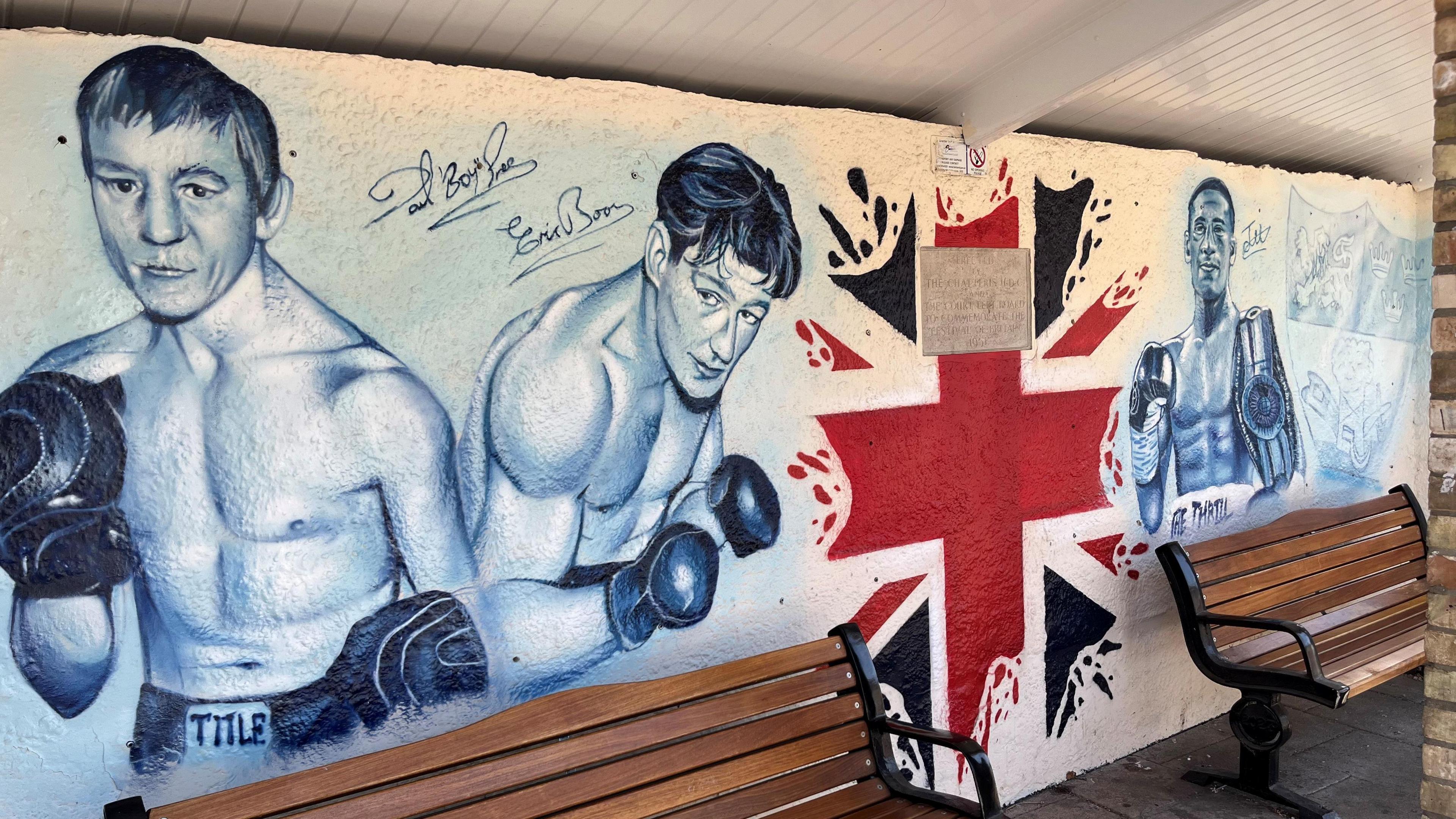 Graffiti art work inside a bus shelter shows three boxers from Chatteris, from left to right, Dave 'Boy' Green, Eric Boon and Jordan Gill. Two wooden benches are in front of the artwork which is done in black and white, with part of a Union flag in the middle.