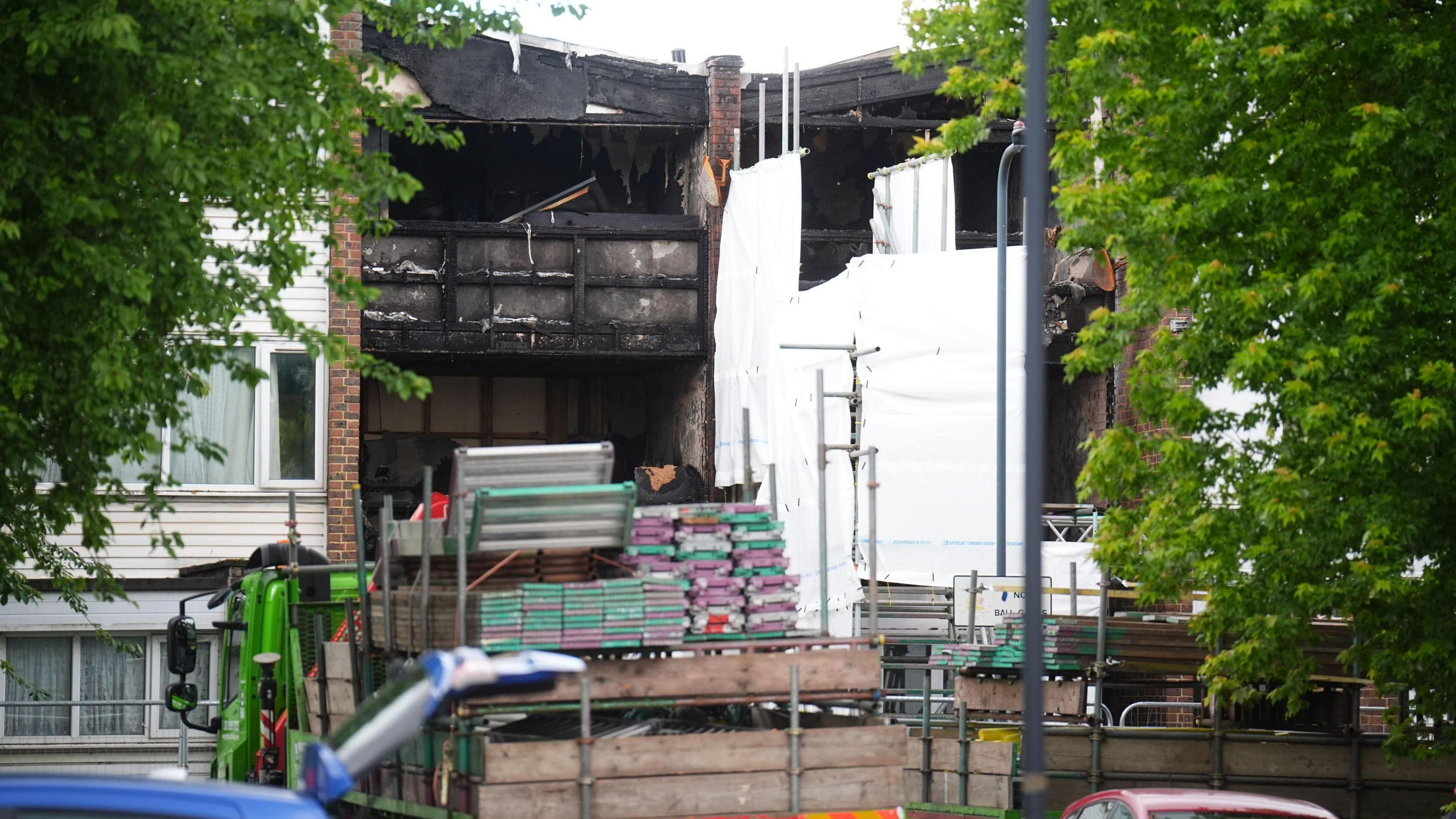 Scaffolding being delivered to the scene of the burned-out houses, which are partially covered with white sheeting