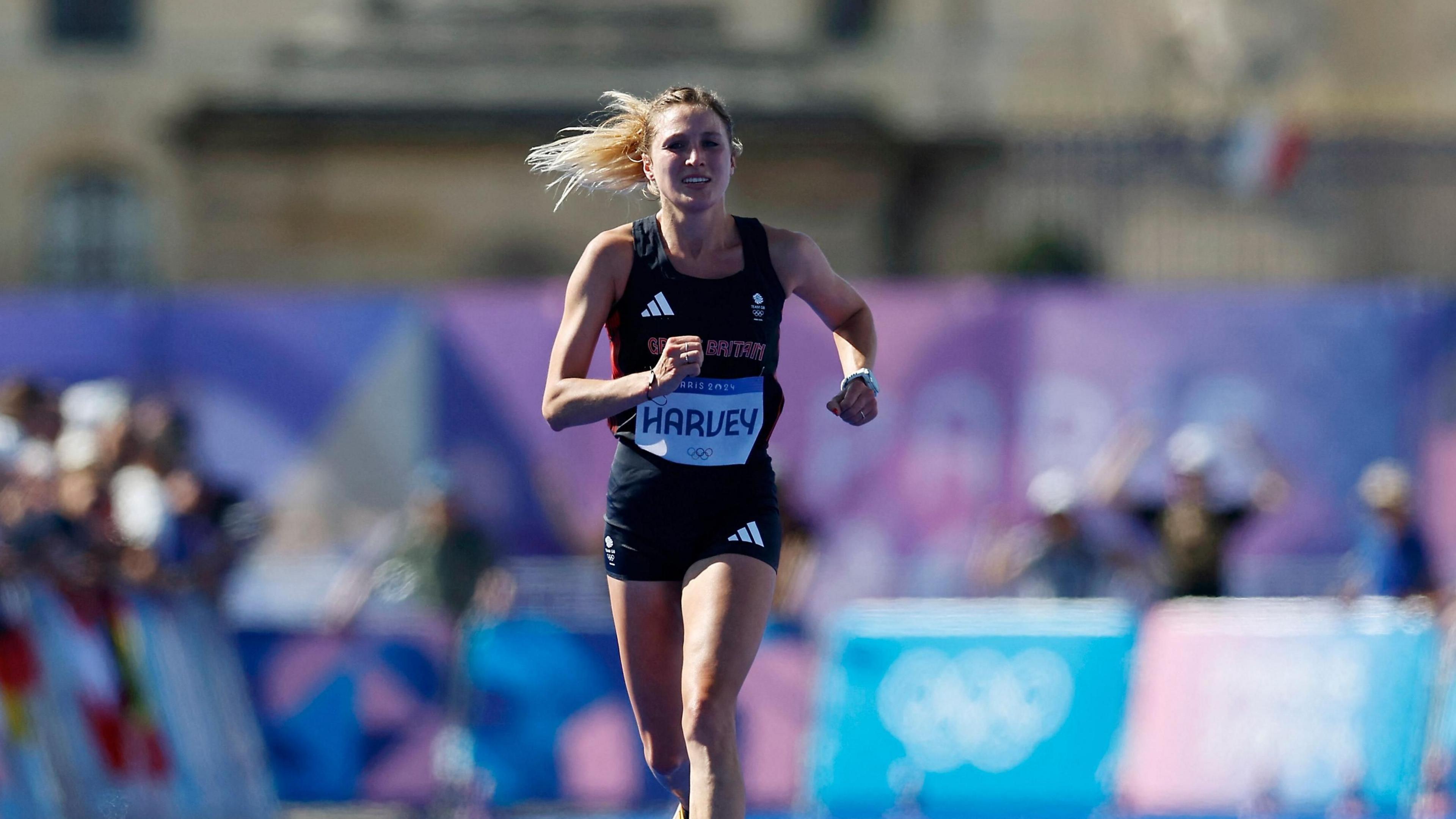 Rose Harvey in black running kit with her tied-back blonde hair flowing behind her against a blurred background of Olympic branding and supporters. 