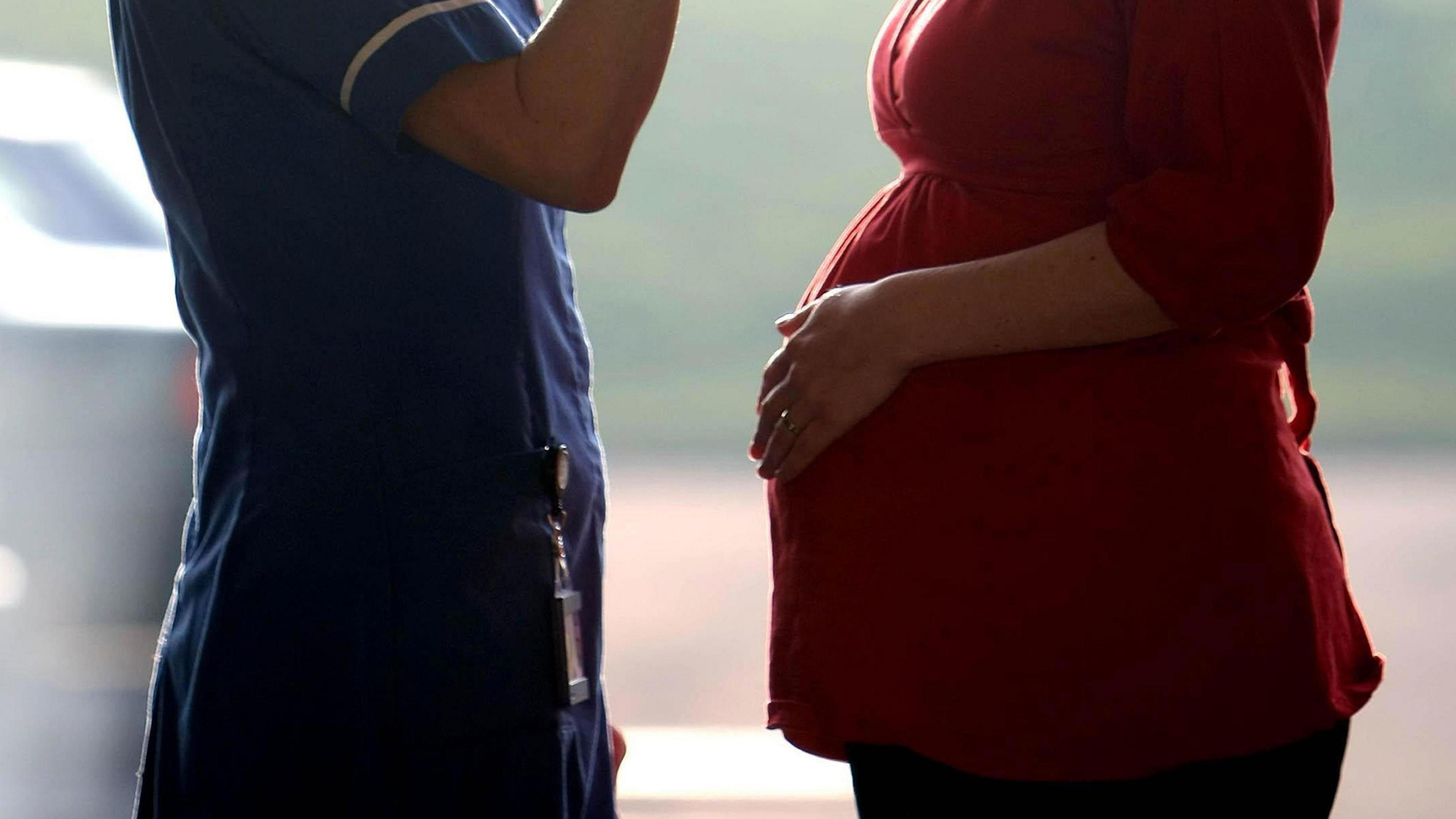 A nurse facing a pregnant woman. Their faces can't be seen. The nurse is wearing a blue uniform and the pregnant women is wearing a red dress.