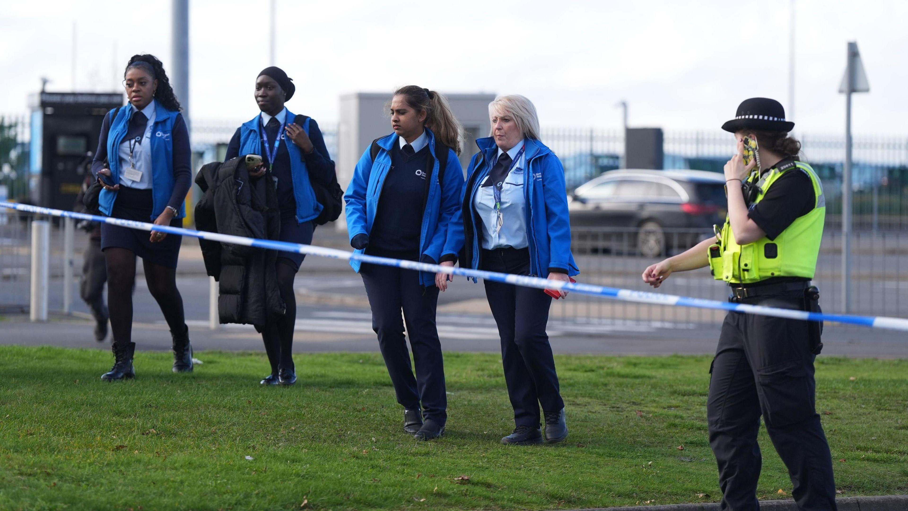 Four women in Birmingham Airport uniform walk towards a police cordon, while a police officer appears to be preparing to lift the tape to let them pass.