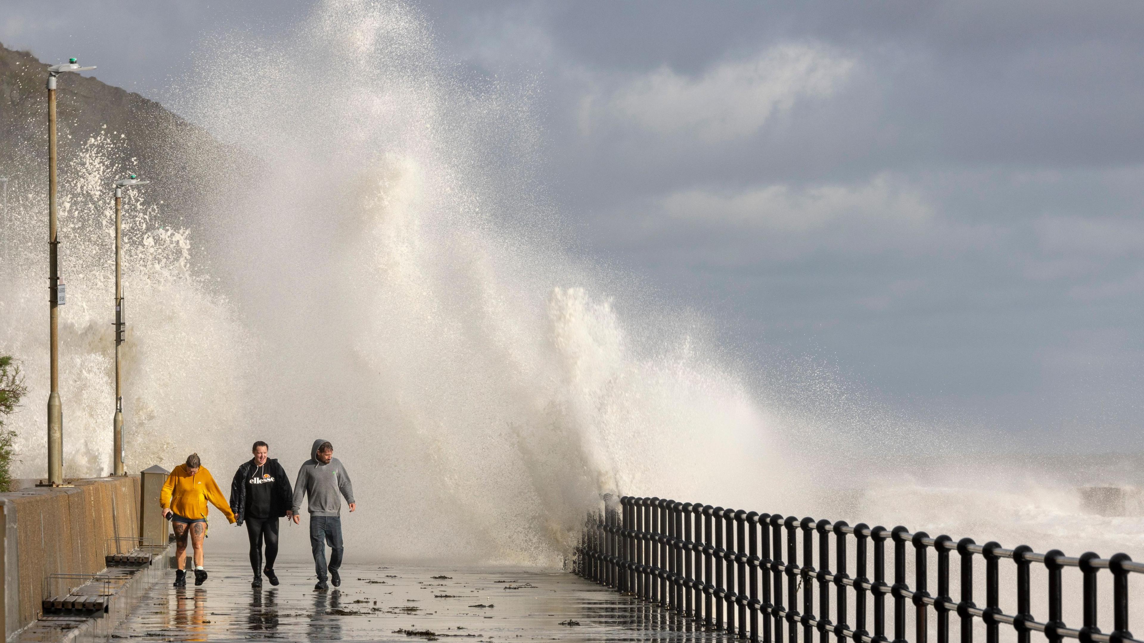 People walk along a promenade while waves crash over behind them