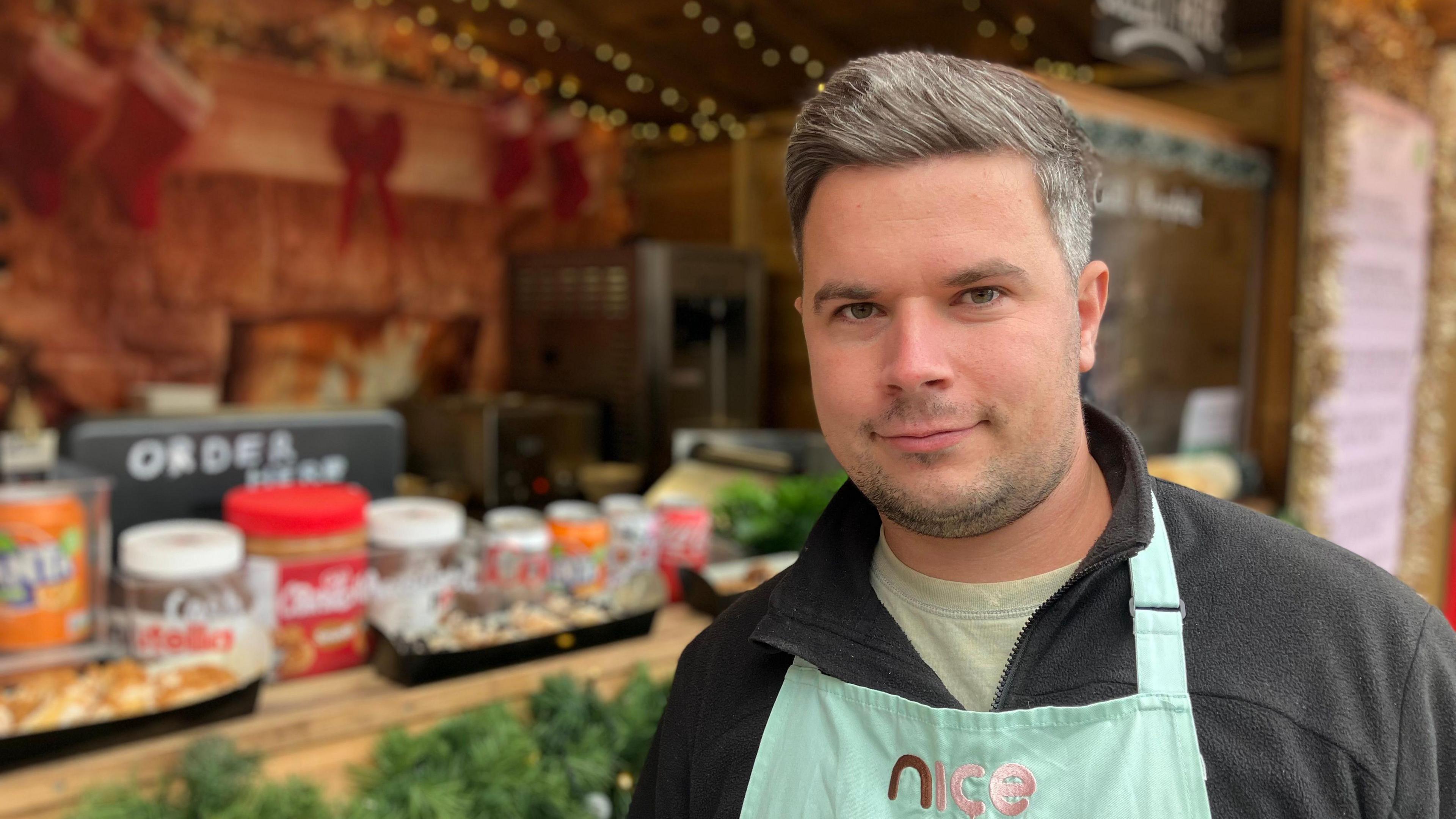 Joseph Malin in a green apron stands in front of a donut stall at a Christmas market. You can see catering equipment and Christmas lights in the background. 