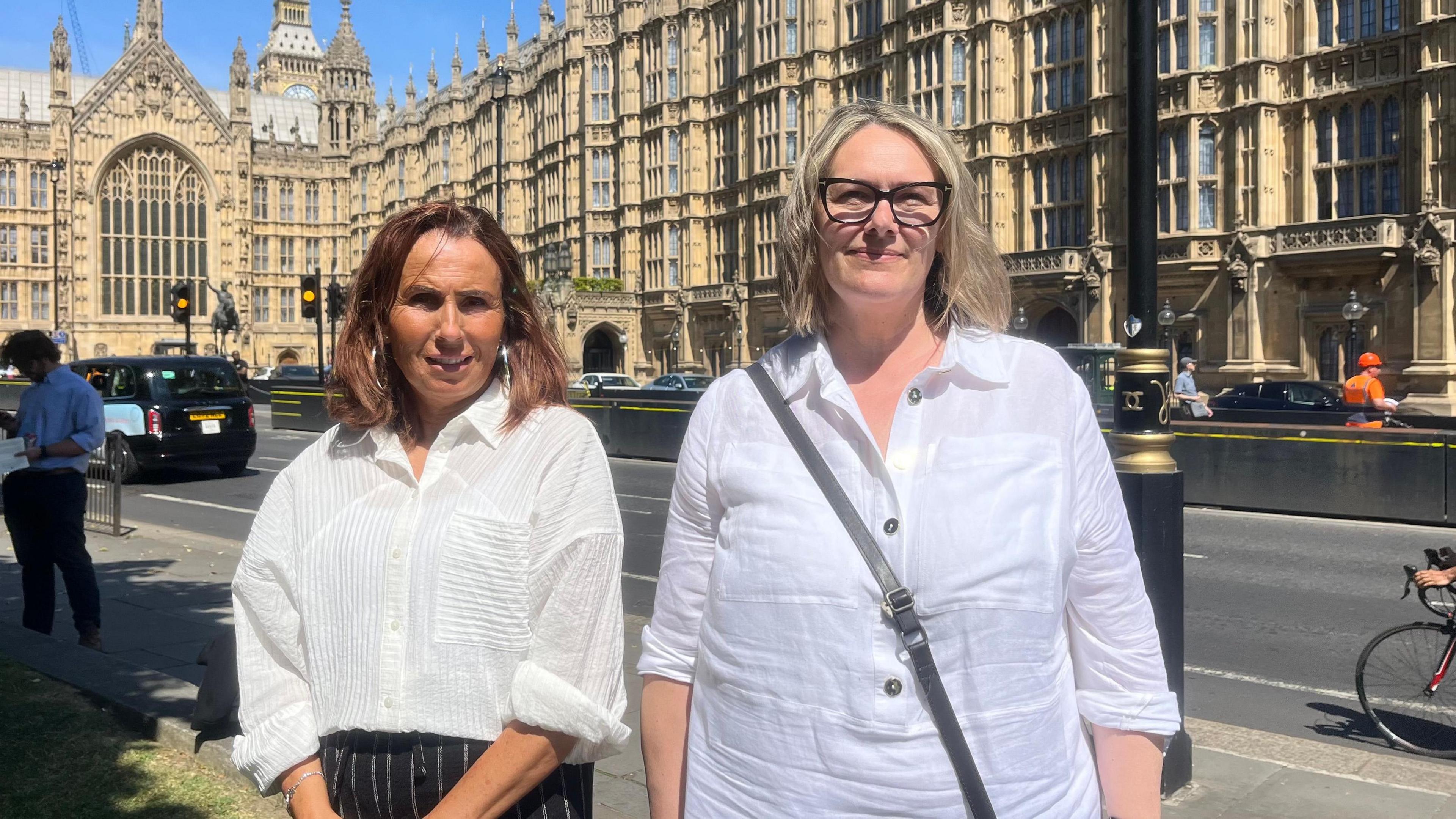 Pictured is Susan Tracey with brown shoulder length hair, a white shirt and black and white stripe trousers. She is standing next to Dawn Style who has blonde shoulder length hair, black big cat eye glasses, an open button white shirt and a black bag strap across her body. They are stood outside Parliament.