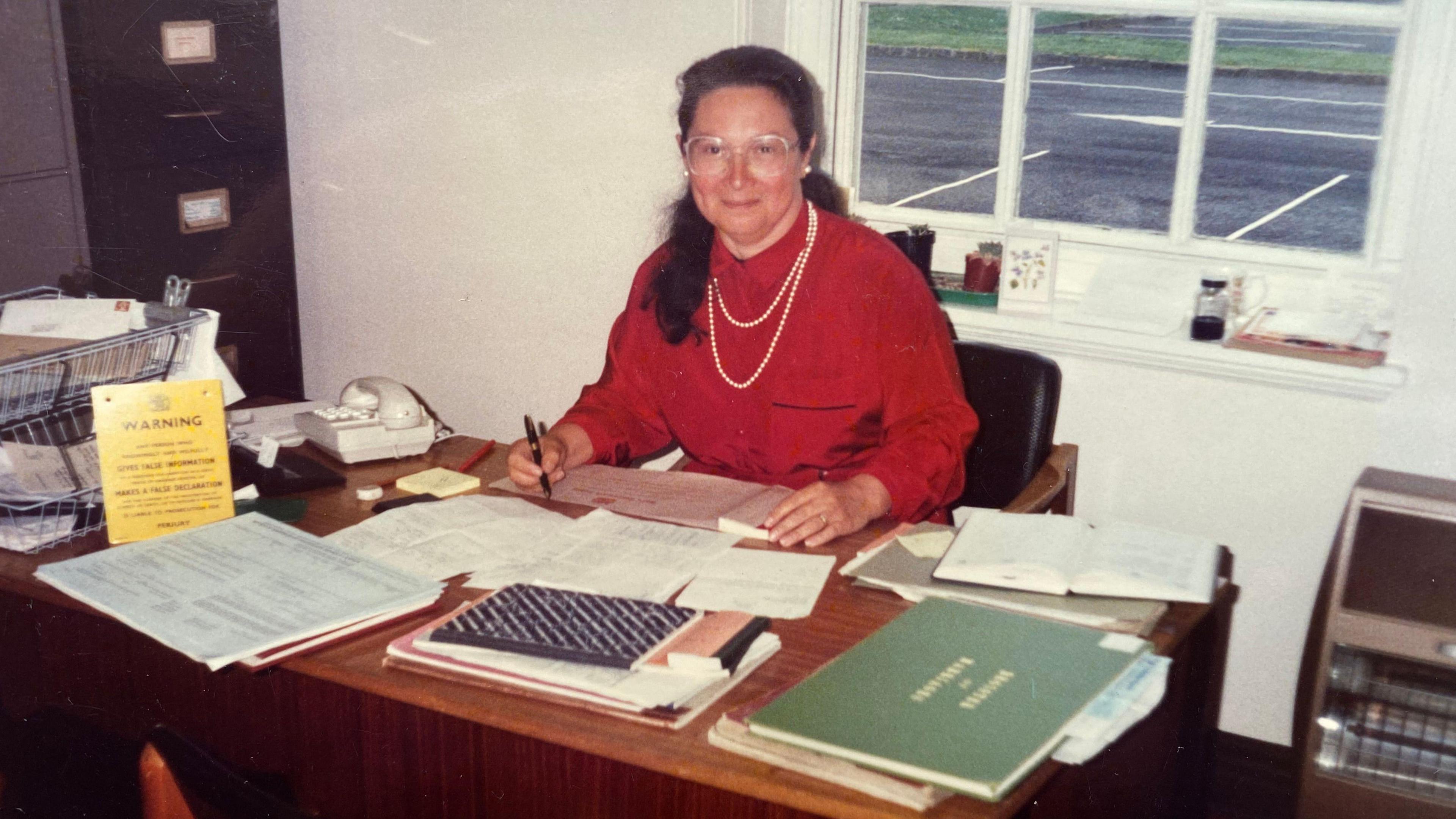 Alicia sits at a wooden desk in a white-walled office. She wears glasses, a red blouse and a necklace of a double string of pearls. The desk is covered with documents, and a filing cabinet can be seen in the corner of the room.