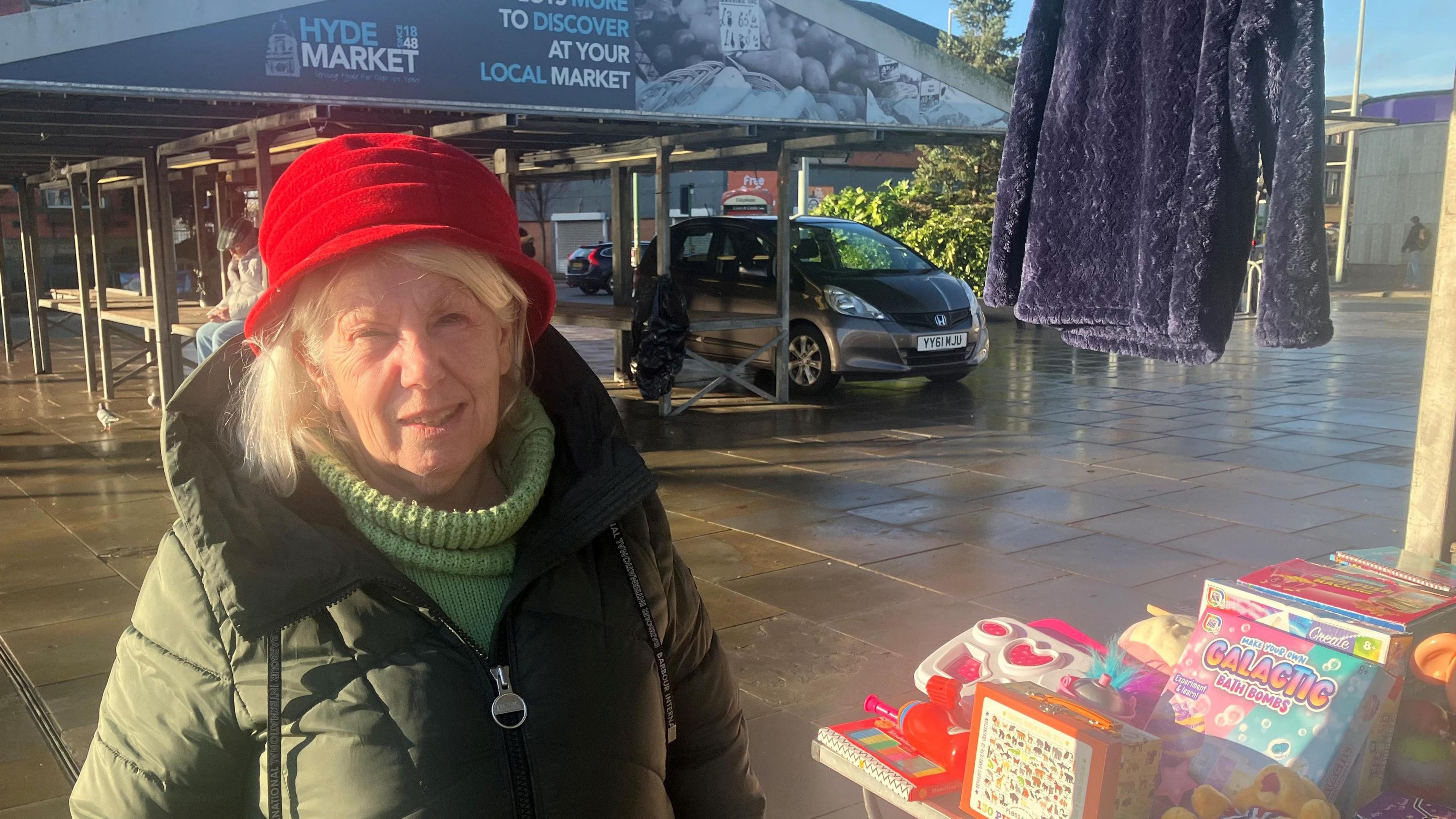 A woman in a red hat, green jumper and coat, stands next to a table full of children's toys and other items underneath a market stand in Hyde.