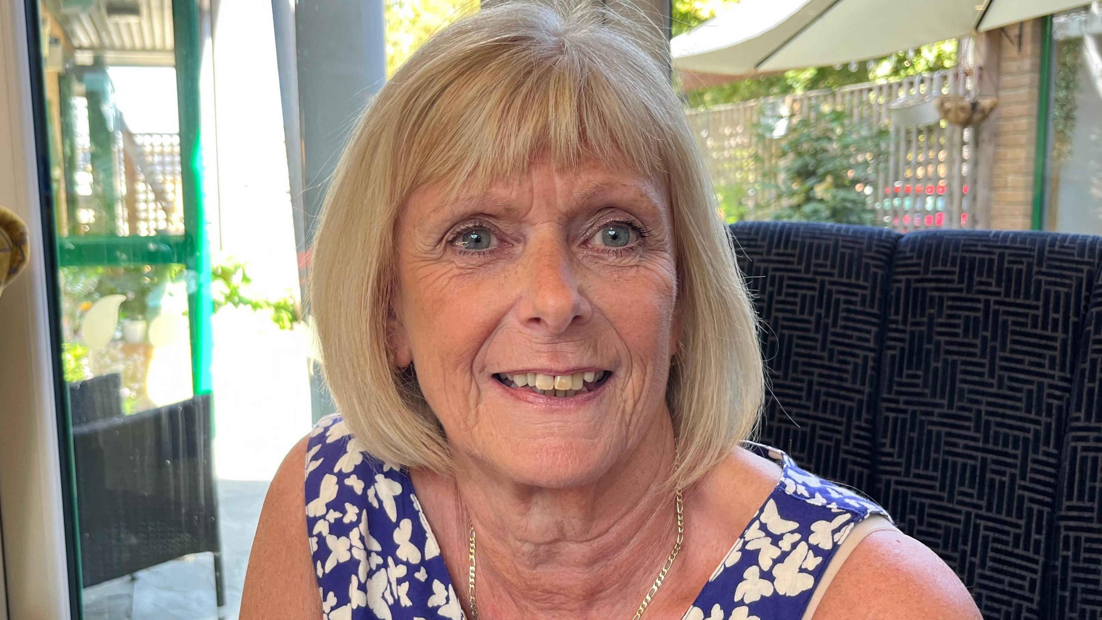 Ms Poole is smiling at the camera. She has medium length blonde hair and blue eyes. She is smiling while sitting inside with light coming in from a window behind her. She is wearing a gold necklace and a blue and white top.