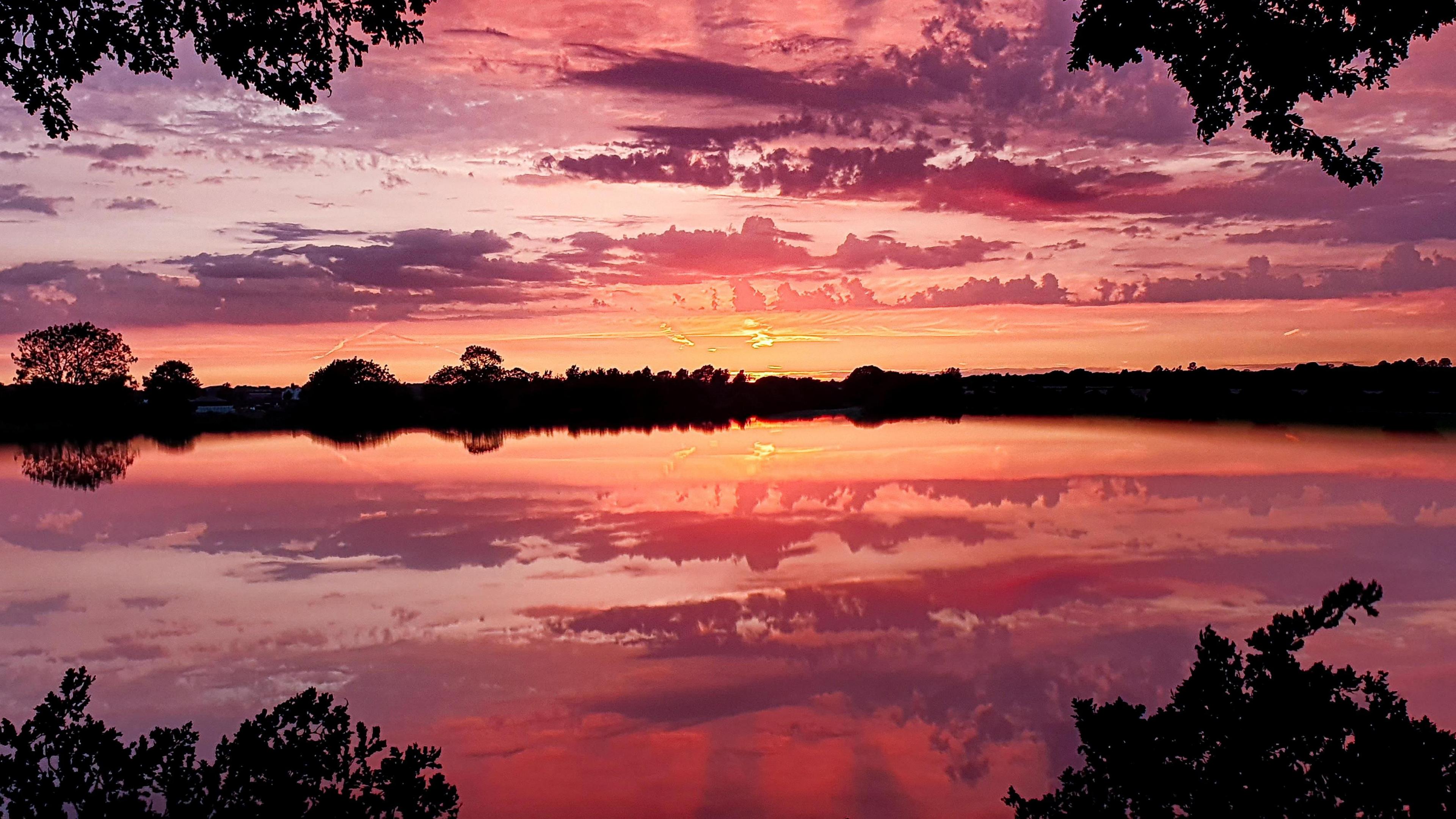 Silhouette of trees in the middle of the picture overlooking a body of still water that has an almost perfect reflection of a purple filled sky created by the sunset with patchy cloud.