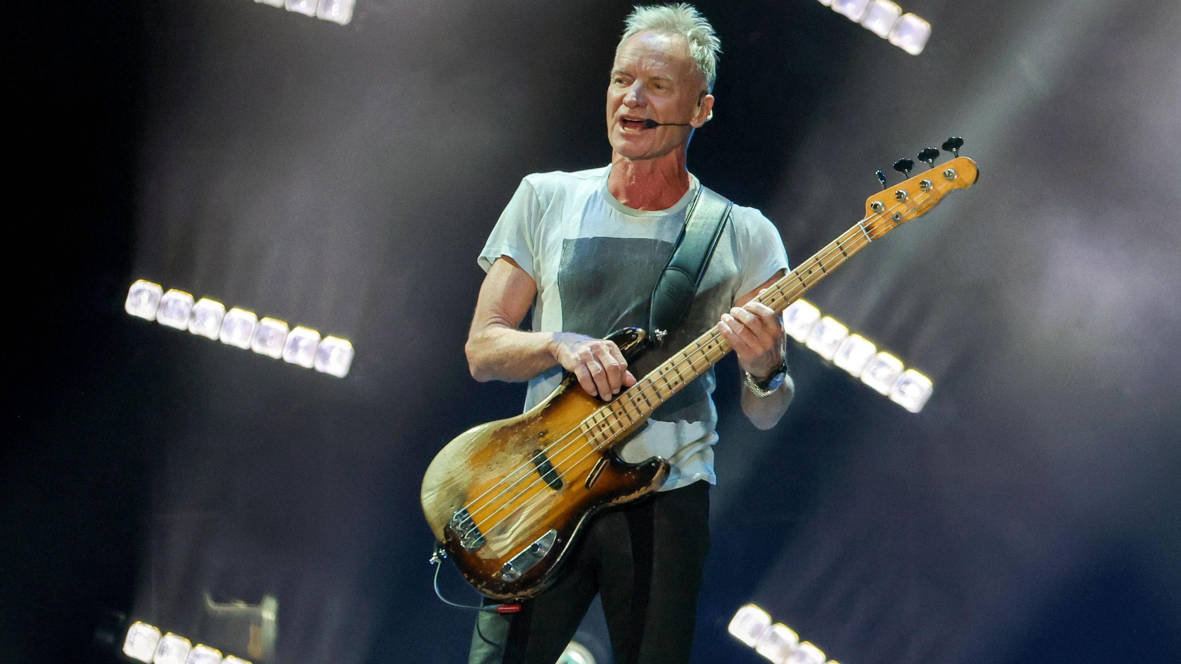 Sting, with spiked fair hair, is on stage wearing a white T-shirt with a black image on the front and is holding an electric guitar.