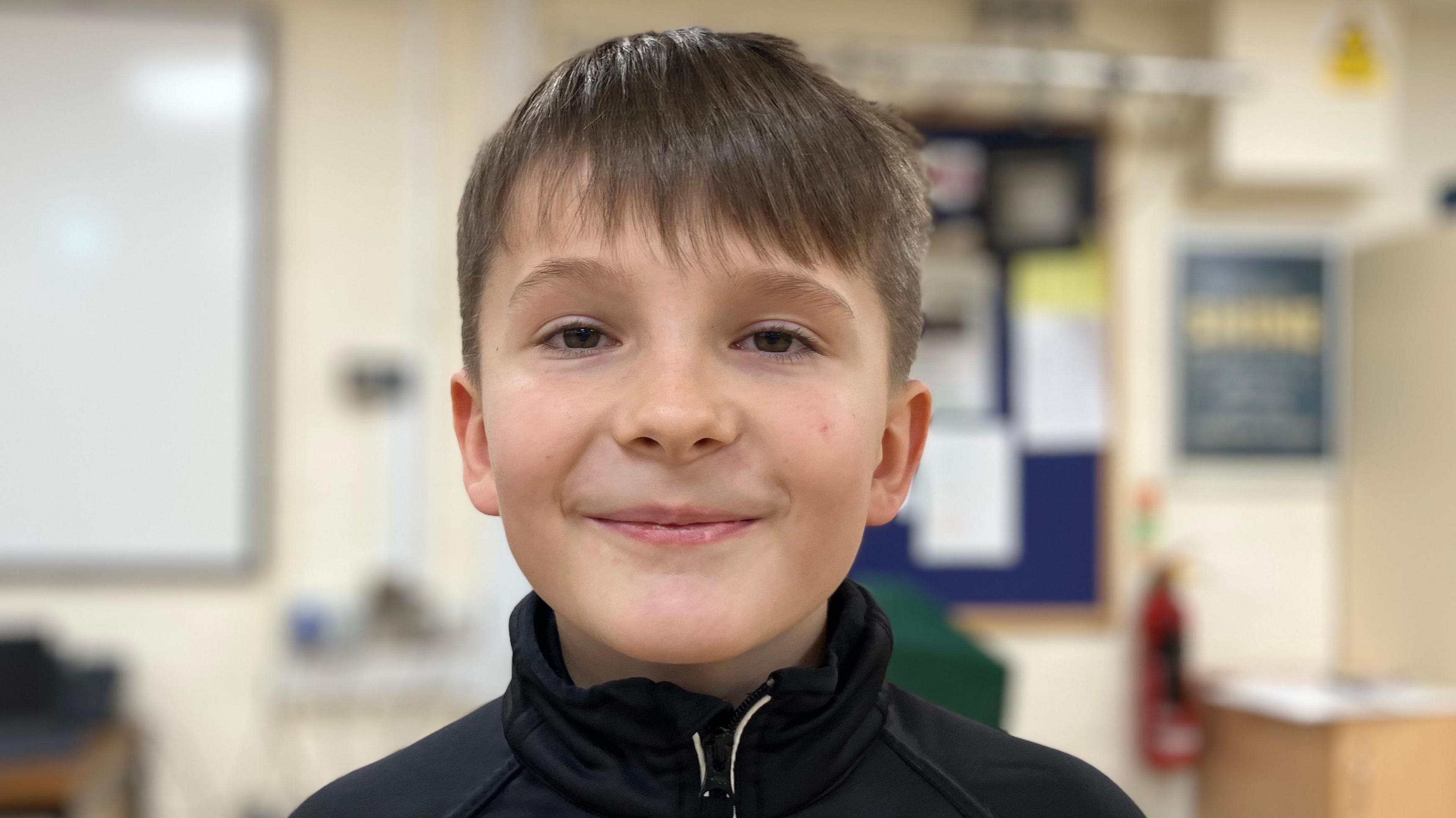 A head and shoulders shot of Blake, a nine year old, smiling and looking at the camera. He is wearing a black top. Behind him is a blurred out wall of a school hall.