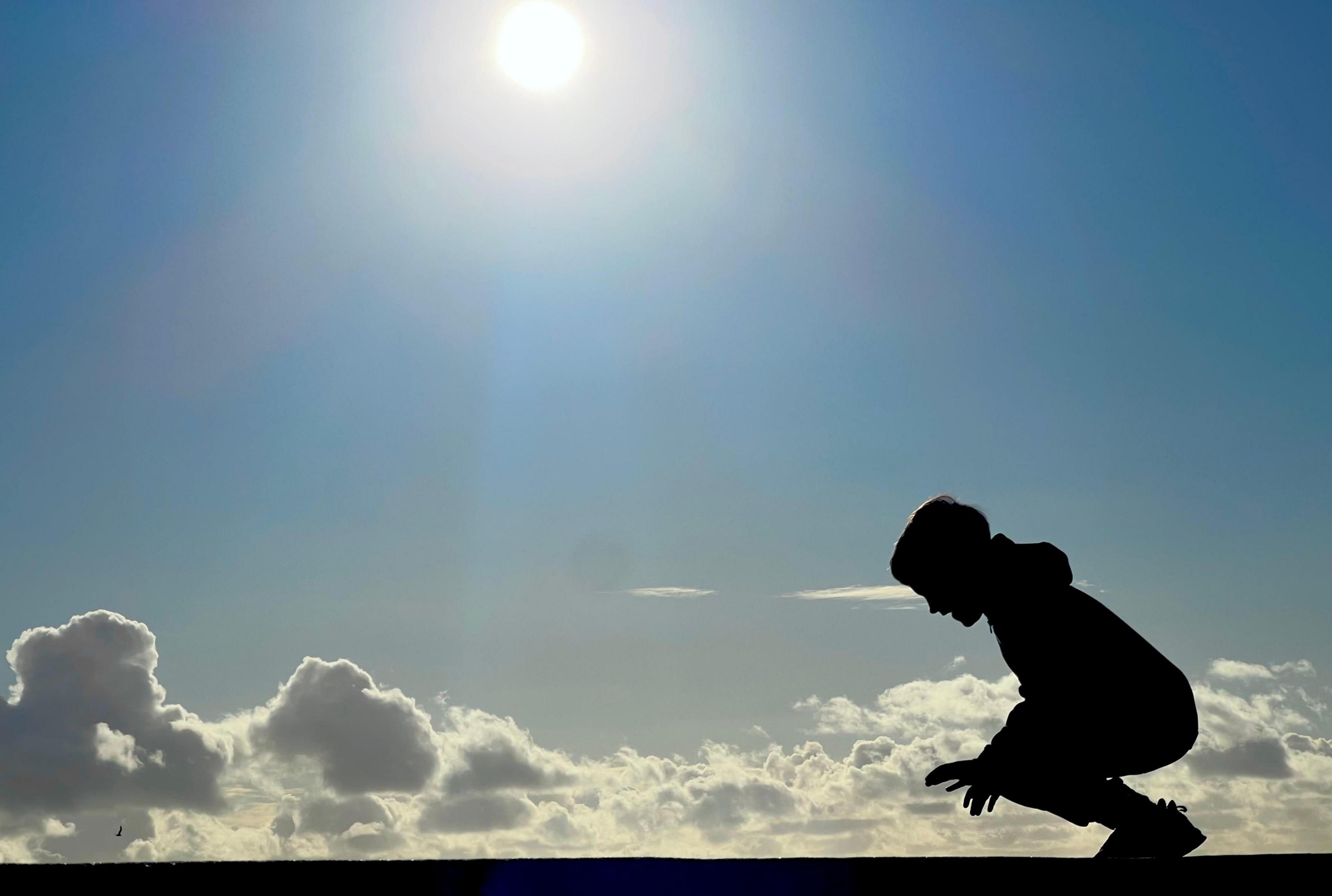 A child crouches down in front of a blue sky and fluffy white clouds. The child is in silhouette. Above the sun is shining brightly. 