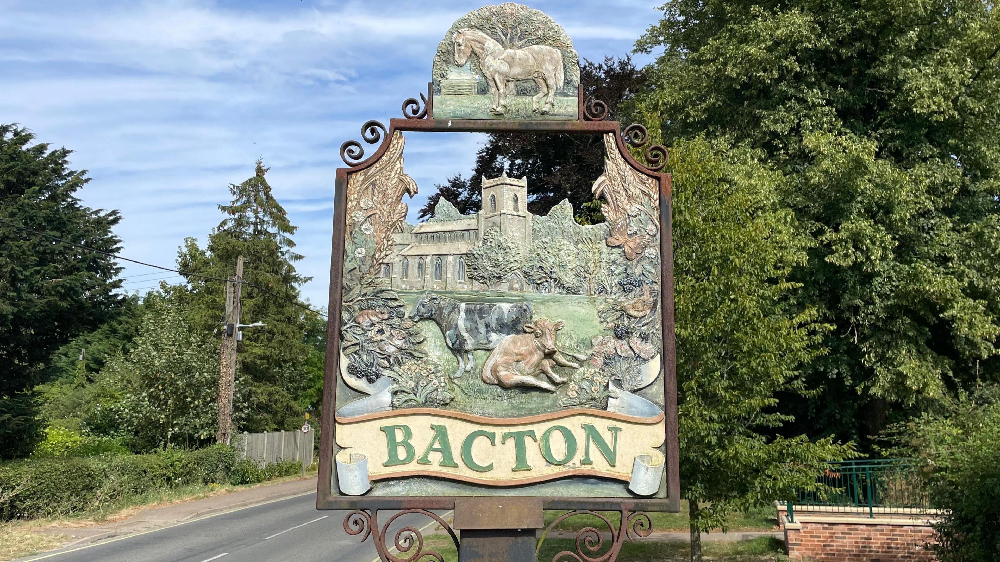 Bacton's village sign showing a horse at the top, a church with trees close by, and cows lying in a grassy field in the foreground. Sheaves of wheat are at the top two corners and agricultural produce around the sides. The word "Bacton" is at the bottom in green lettering on a yellow ribbon.