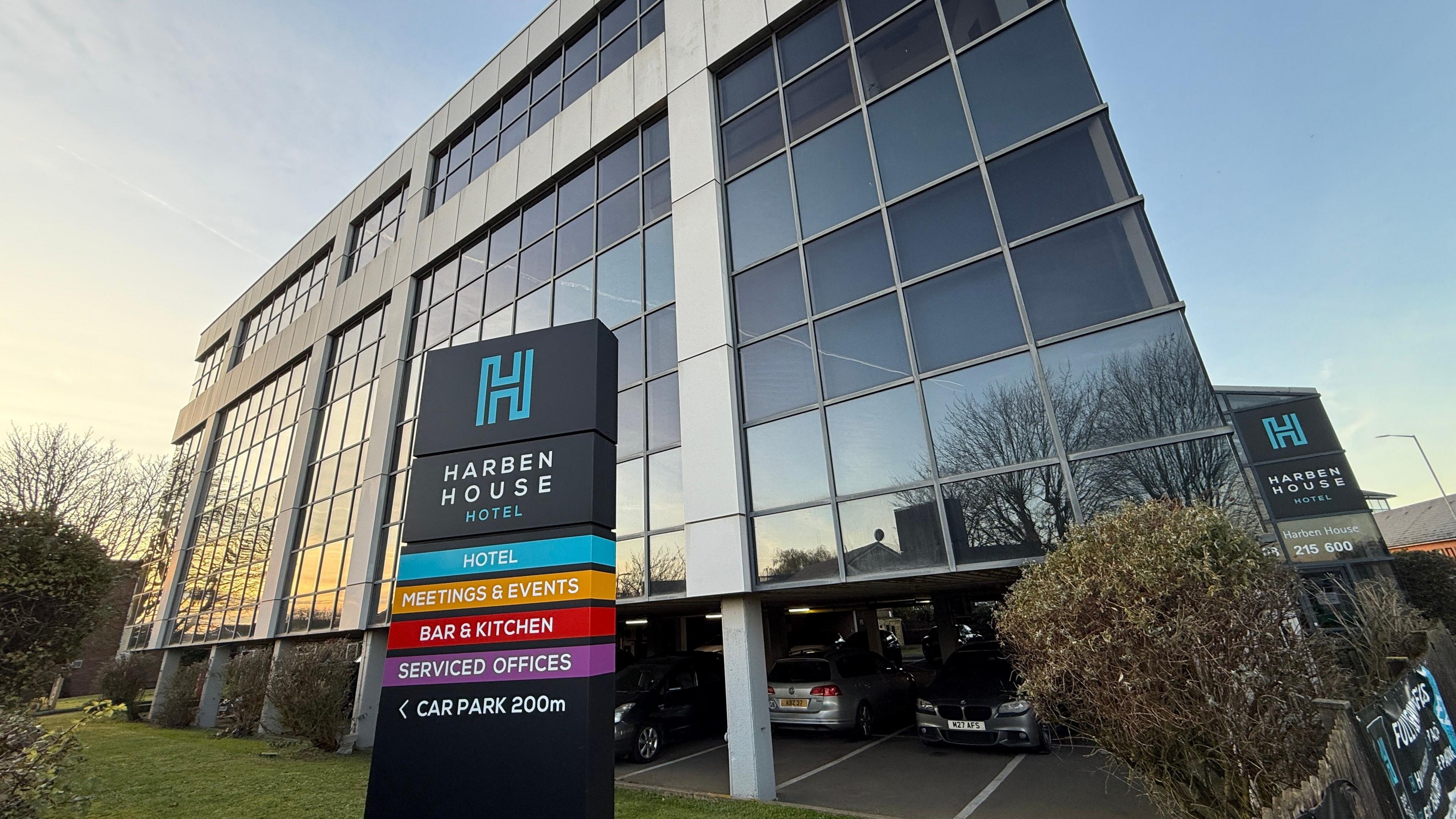 Exterior shot of Harben House Hotel, a mainly glass angular building with a sign in front of it. In front of a blue sky. There is bush to the right of the picture.