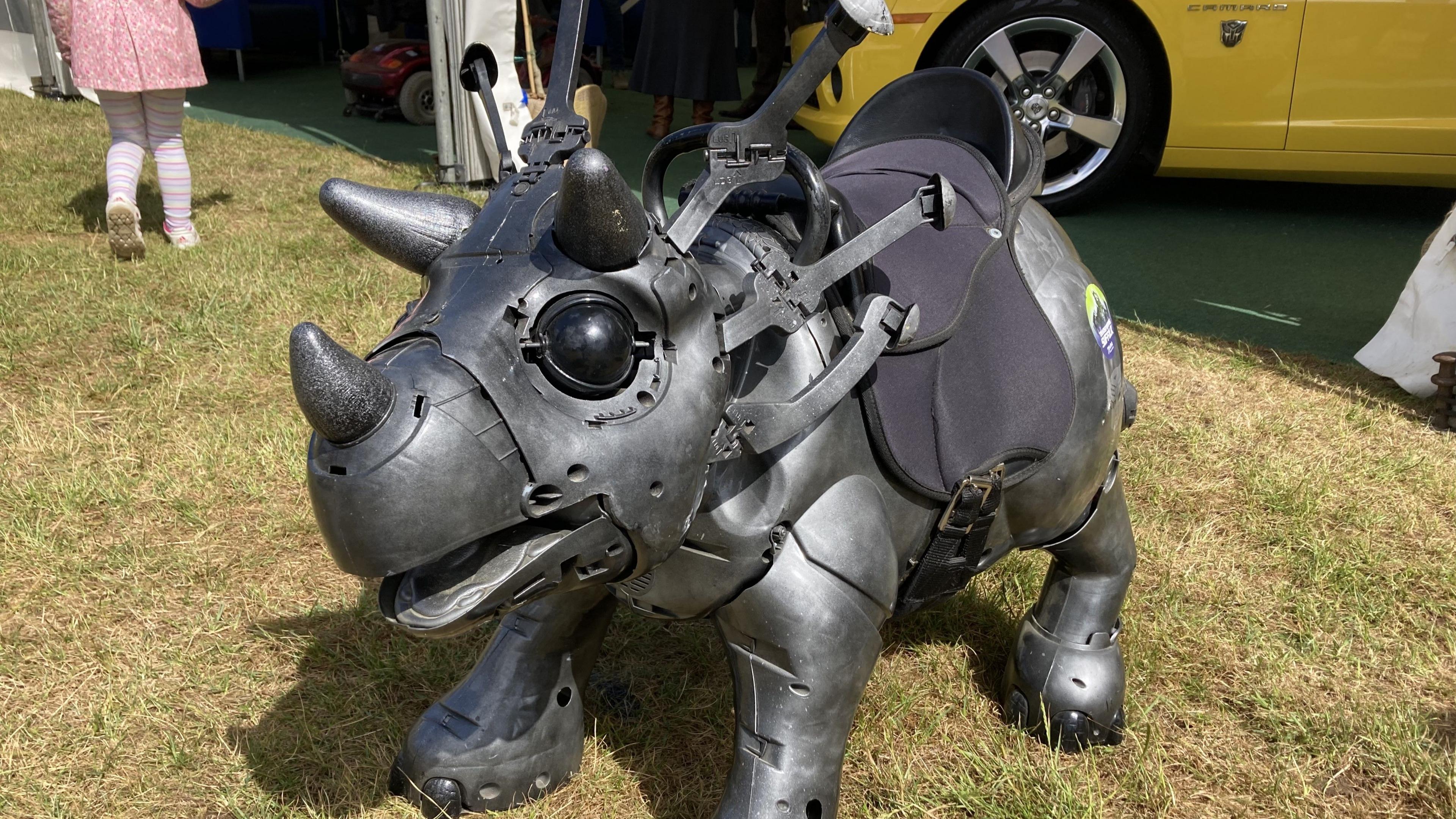A robotic looking dinosaur stands in a grass field. It is a dark grey colour and has three horns on its head. It has a black saddle fitted to its body.