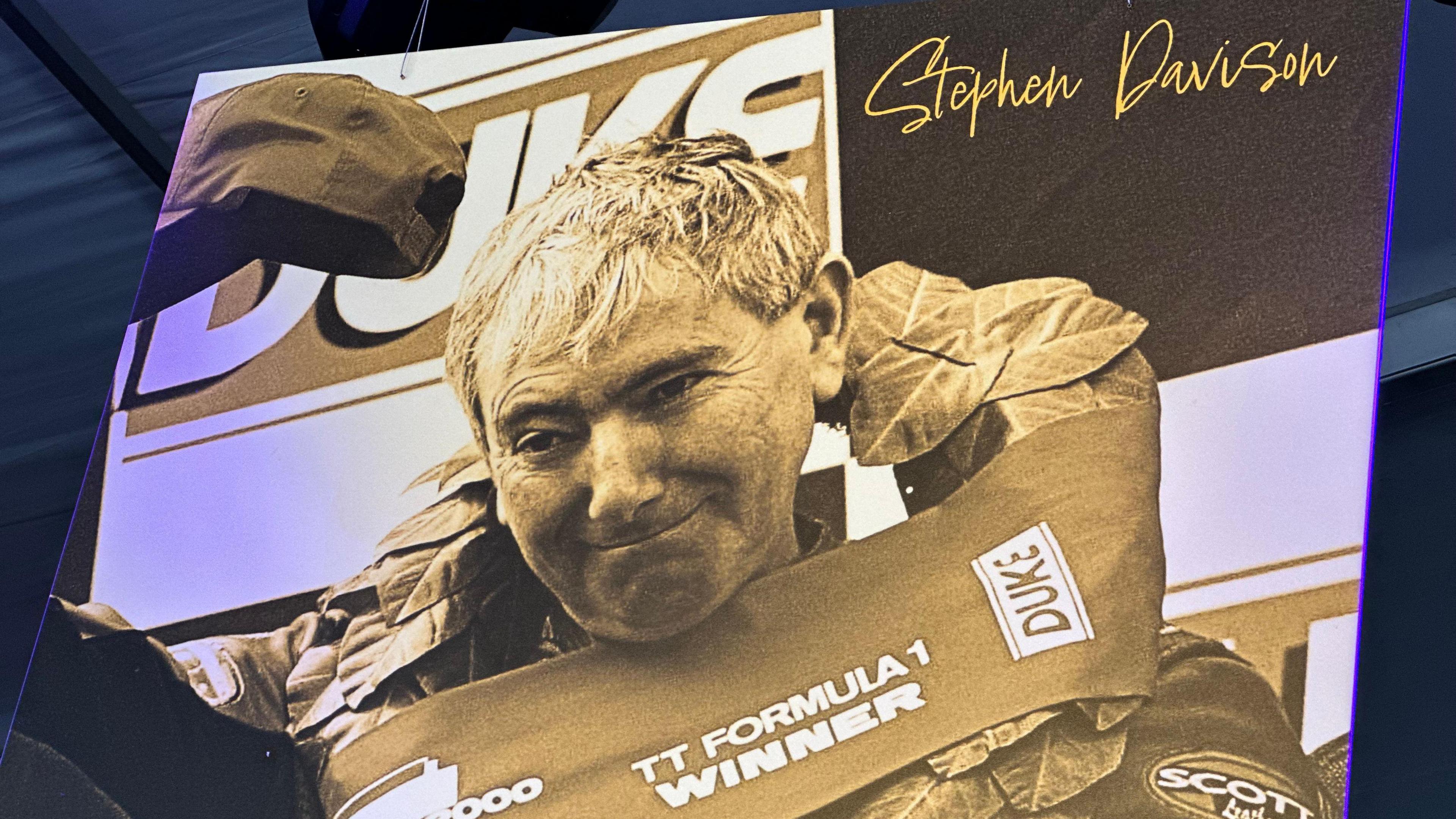 A close-up of a large sepia toned photo of Joey Dunlop which is on display at the exhibition. It shows him raising his cap in celebration on the podium in what was his final TT win. He has a laurel wreath around his neck which reads TT Formula 1 winner.