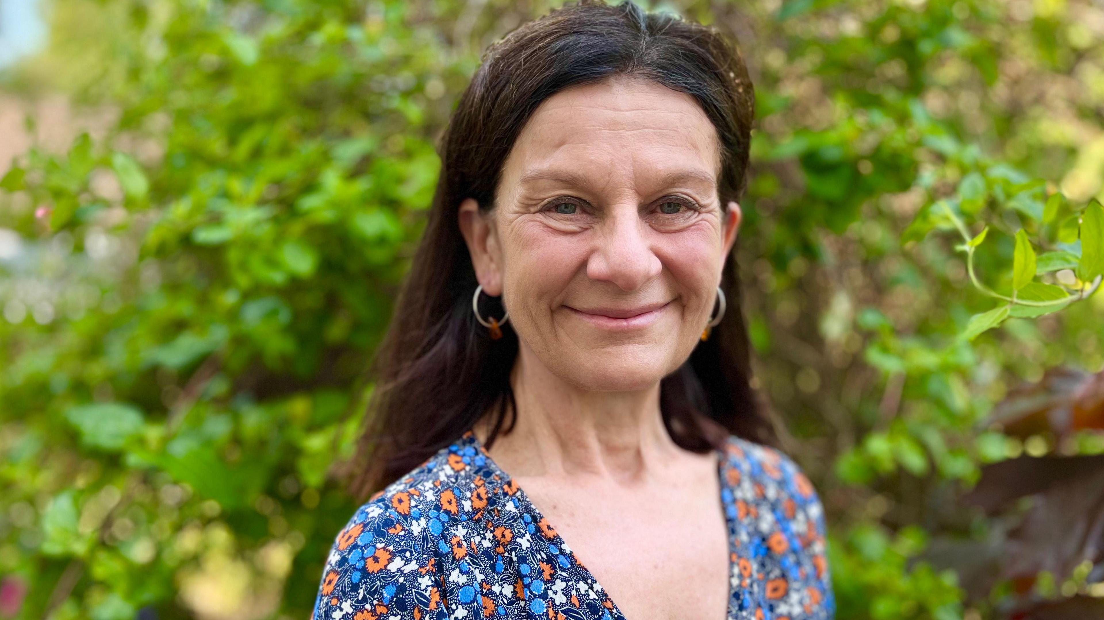 Bridget Smith, who has long dark hair falling down her back . She is wearing a floral top in blue, orange and white and is smiling towards the camera