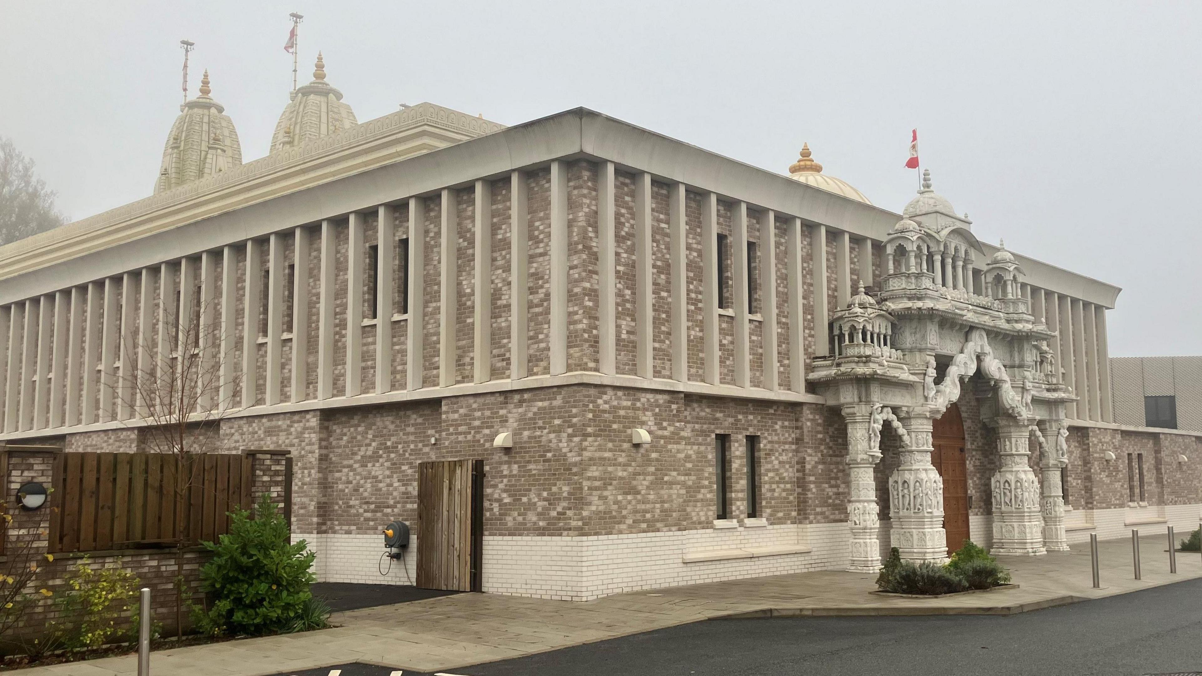 A large pale brick temple with a decorative entrance and has towers at the back. Silver bollards are out the front.