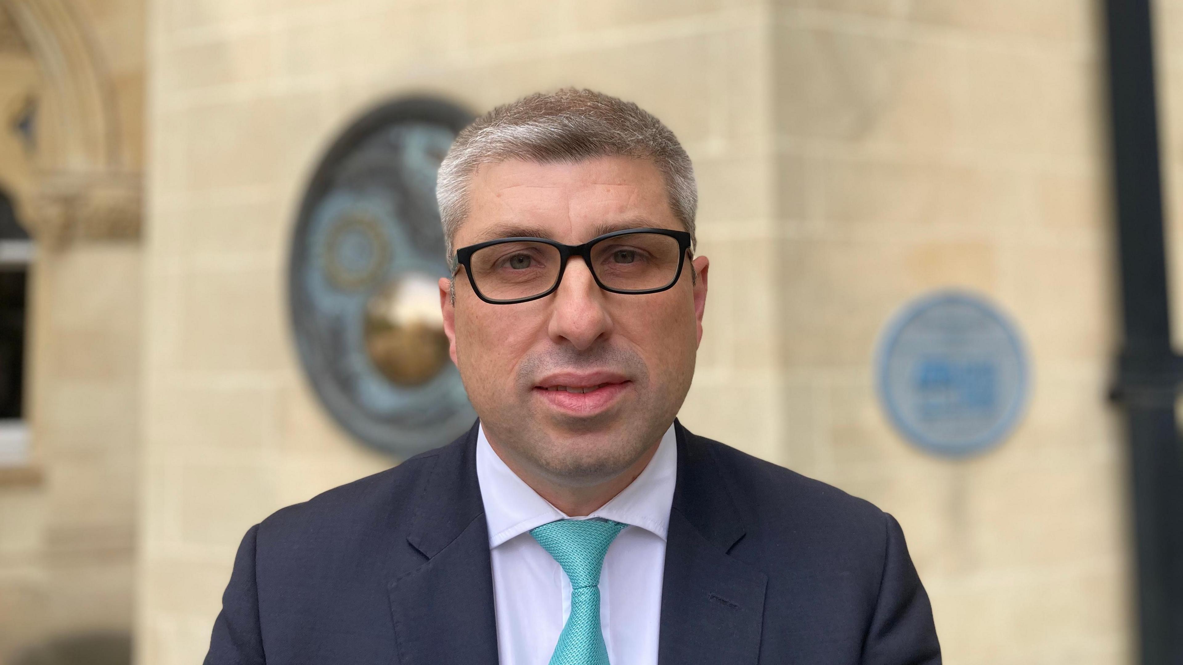 Mark Arnull with short dark hair and black-framed glasses wearing a light-coloured shirt, green tie and dark jacket. He is looking at the camera and is standing in front of the light stone wall of Northampton Guildhall