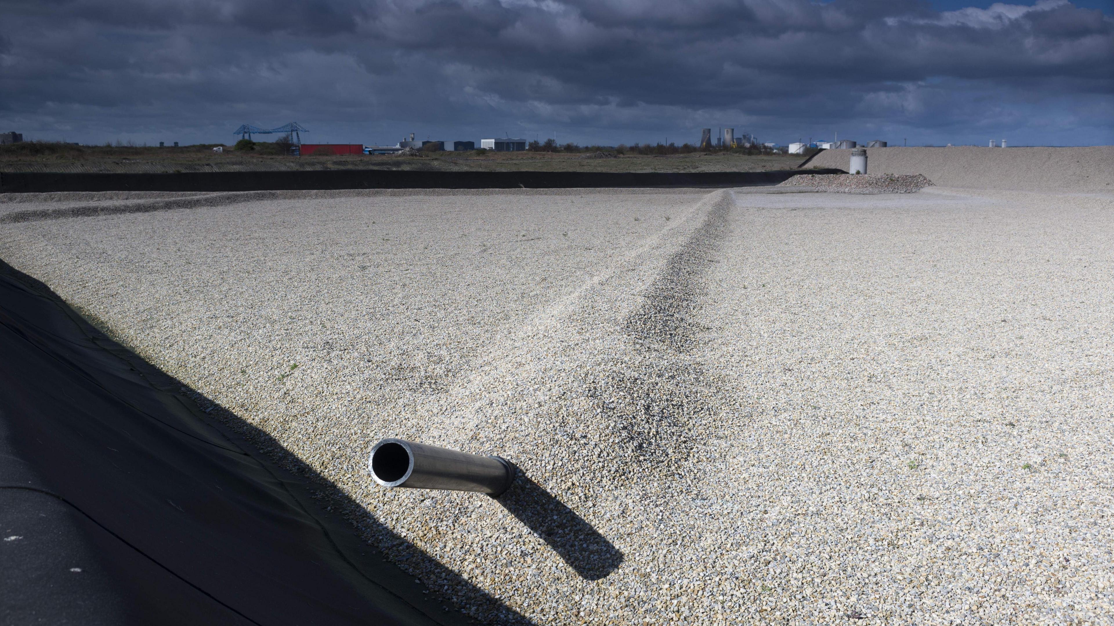 A newly engineered landfill cell with a silver pipe emerging from the gravel. The industrial landscape can be seen in the background. 