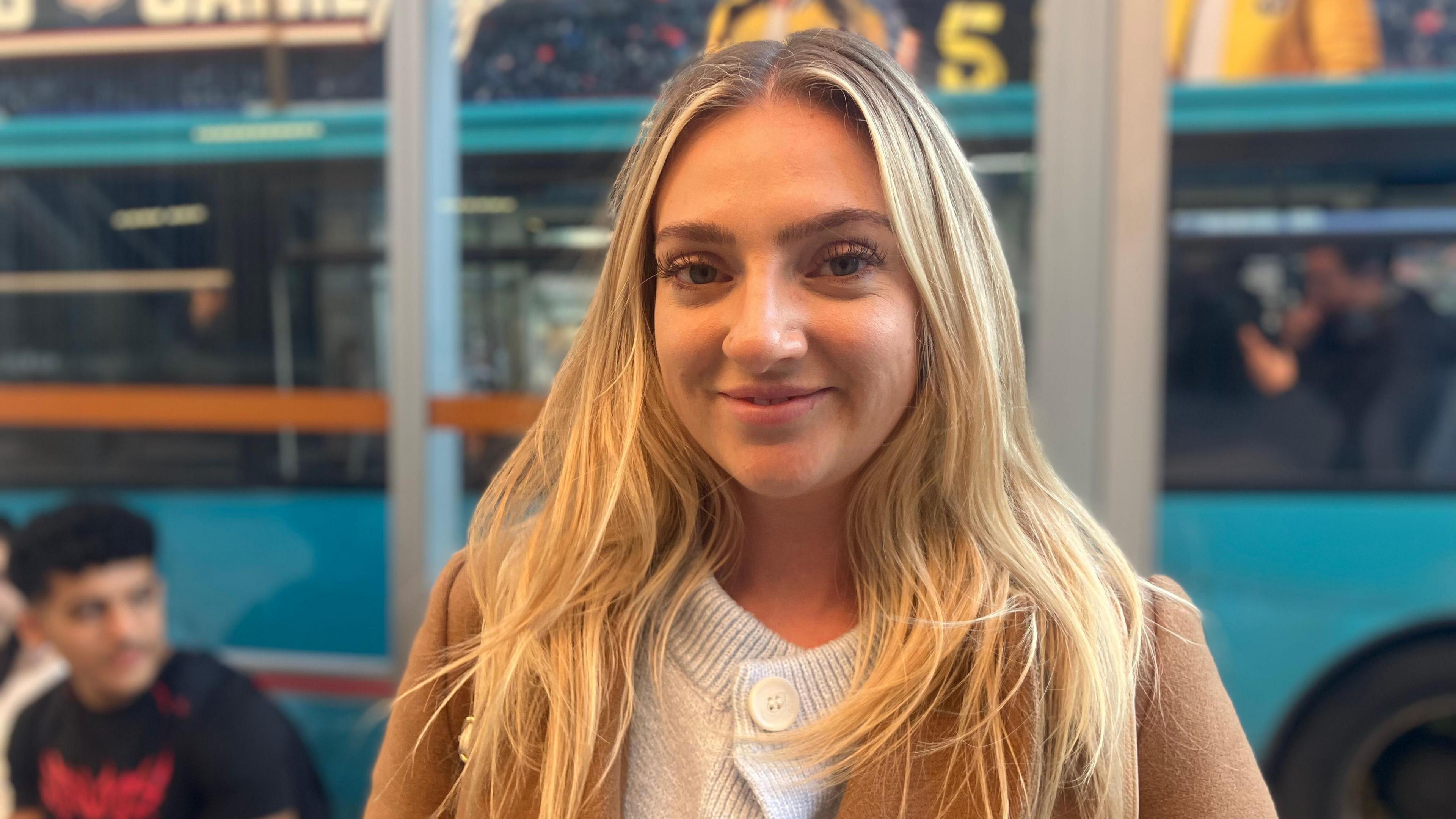 A woman with blonde hair stands in front of a bus stop. Behind her, a teal bus is parked up and blurred. She wears an oatmeal coloured cardigan and a camel coat.