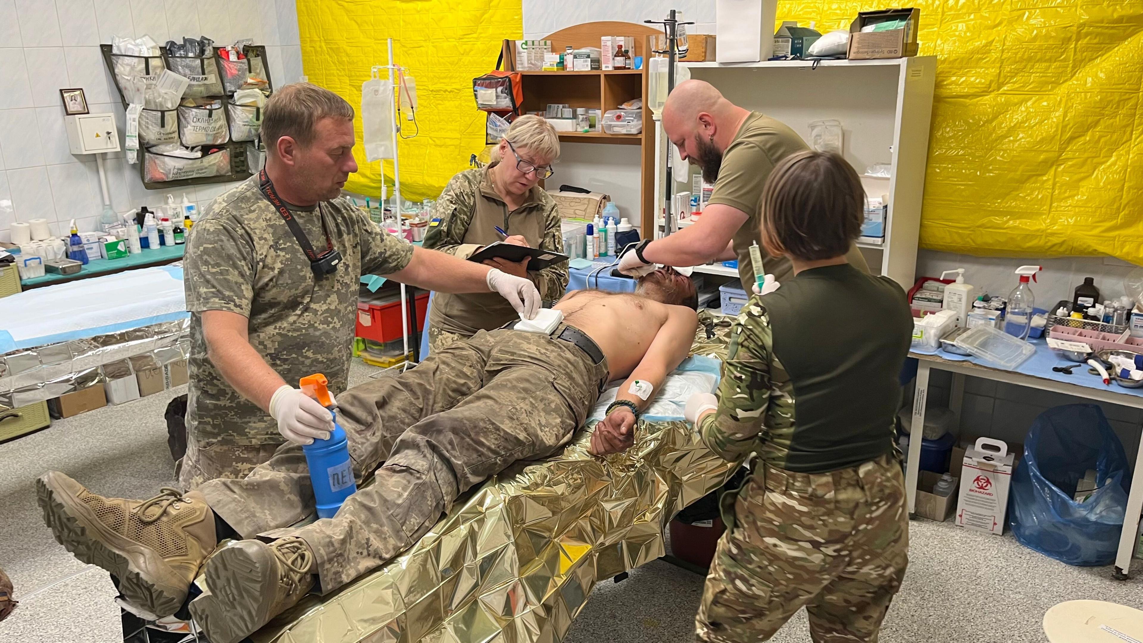 Medics treat a soldier injured in the battle for Pokrovsk. The soldier is laid out on a treatment table - naked from the waist up. Four medics, two men and two women, are treating him. There are two big plastic yellow sheets up at the two windows.