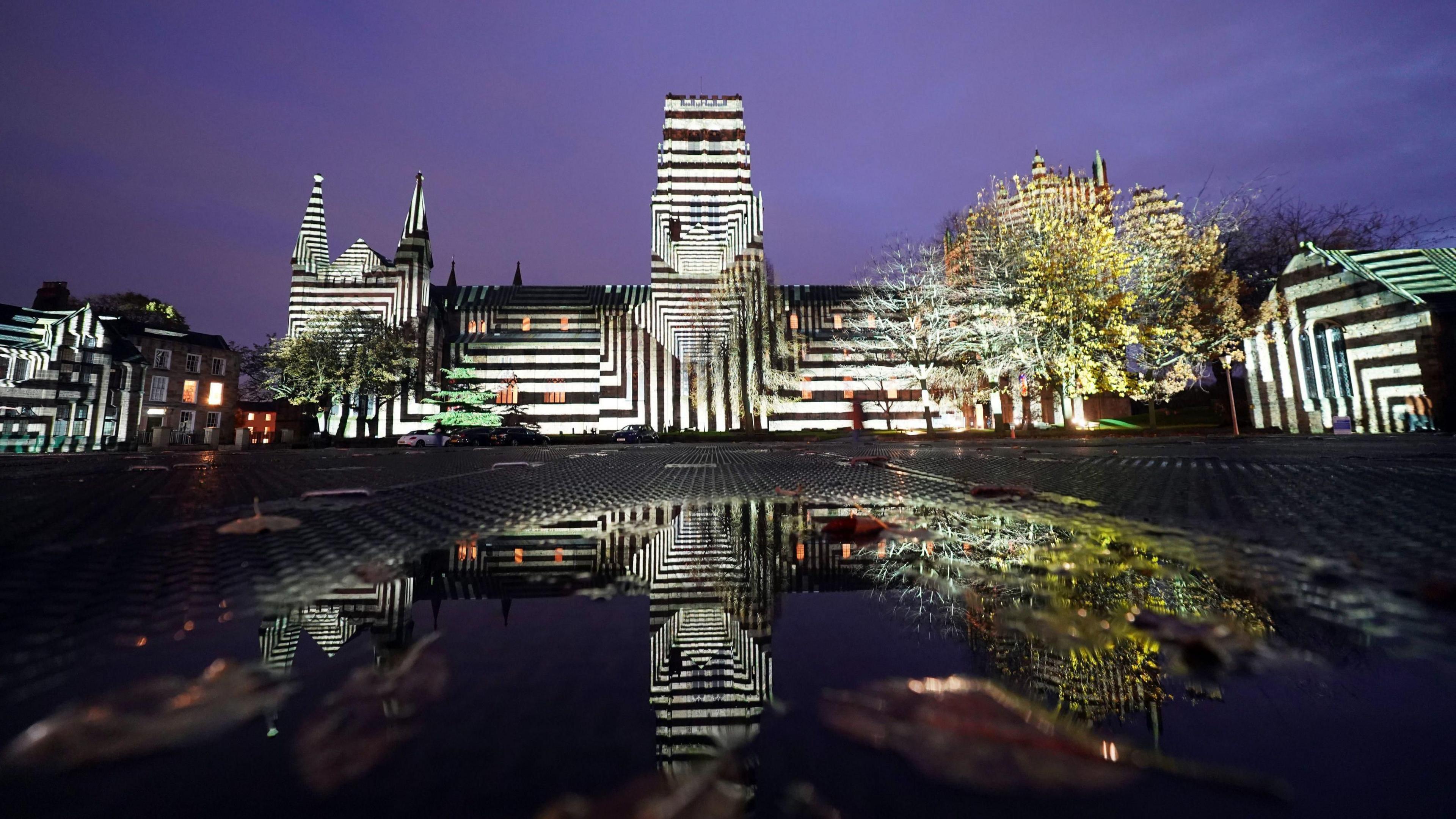 'Liquid Geomotry' lights up Durham Cathedral during a preview for Lumiere 2023. The sprawling medieval stone building is illuminated in black and white vertical and horizontal stripes, reminiscent of a geometric zebra. The building and lights are reflected in water in the foreground.