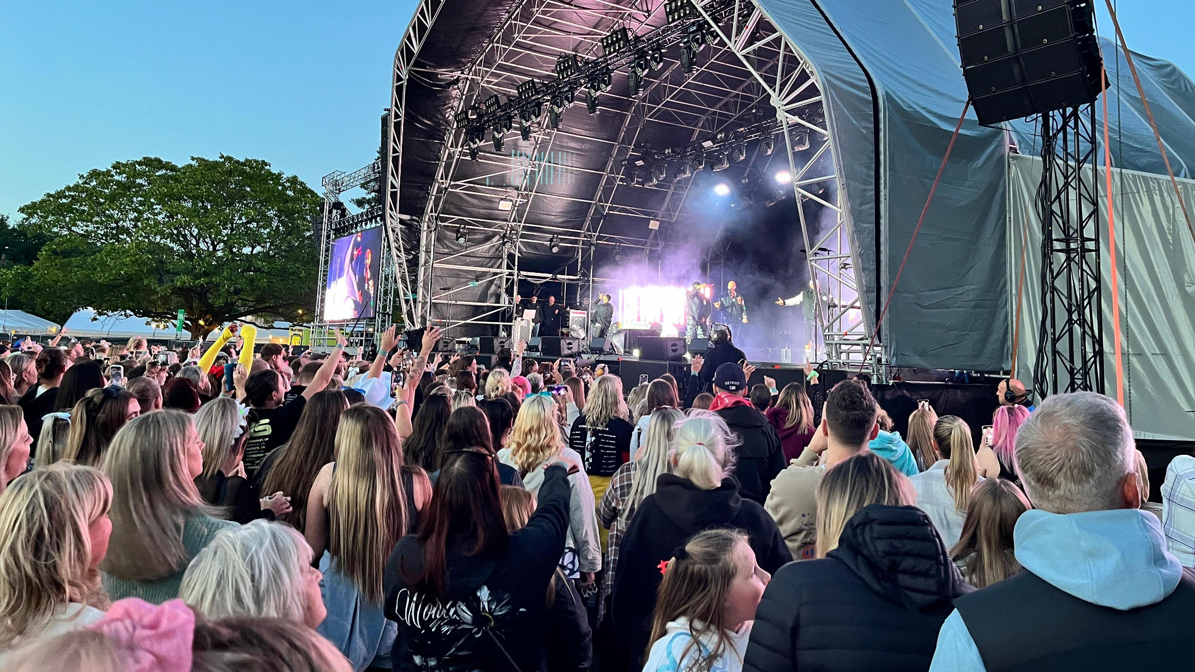 A stage with 4 men, JLS, performing on, with lights. The sky is an inky blue. The crowd stretches all the way from the stage and back past the photographer. There are mature trees in the distance.