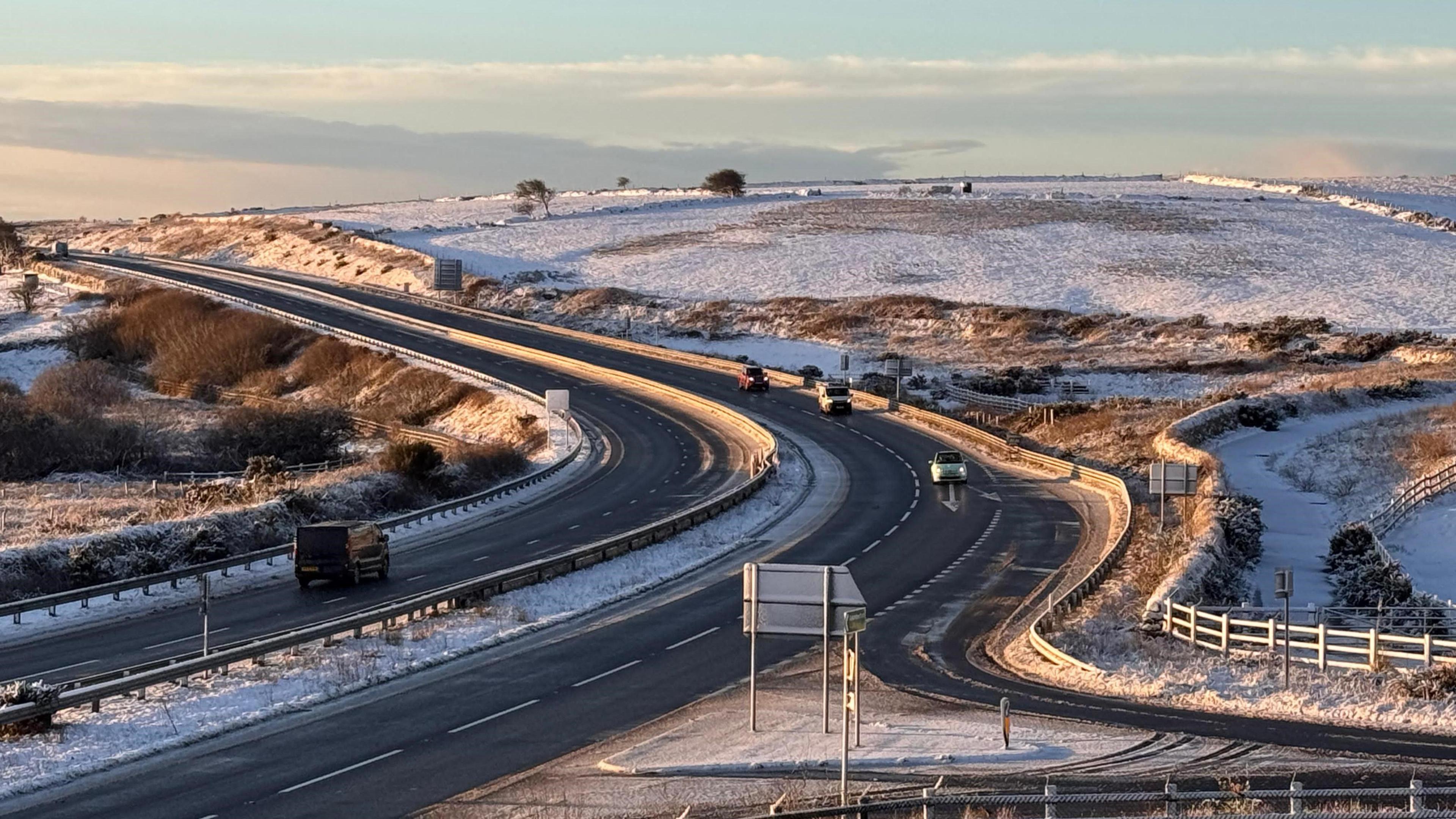 The A30 at Temple, Bodmin is surrounded by snowy fields. There are very few cars on the road and there is a pale blue sky.