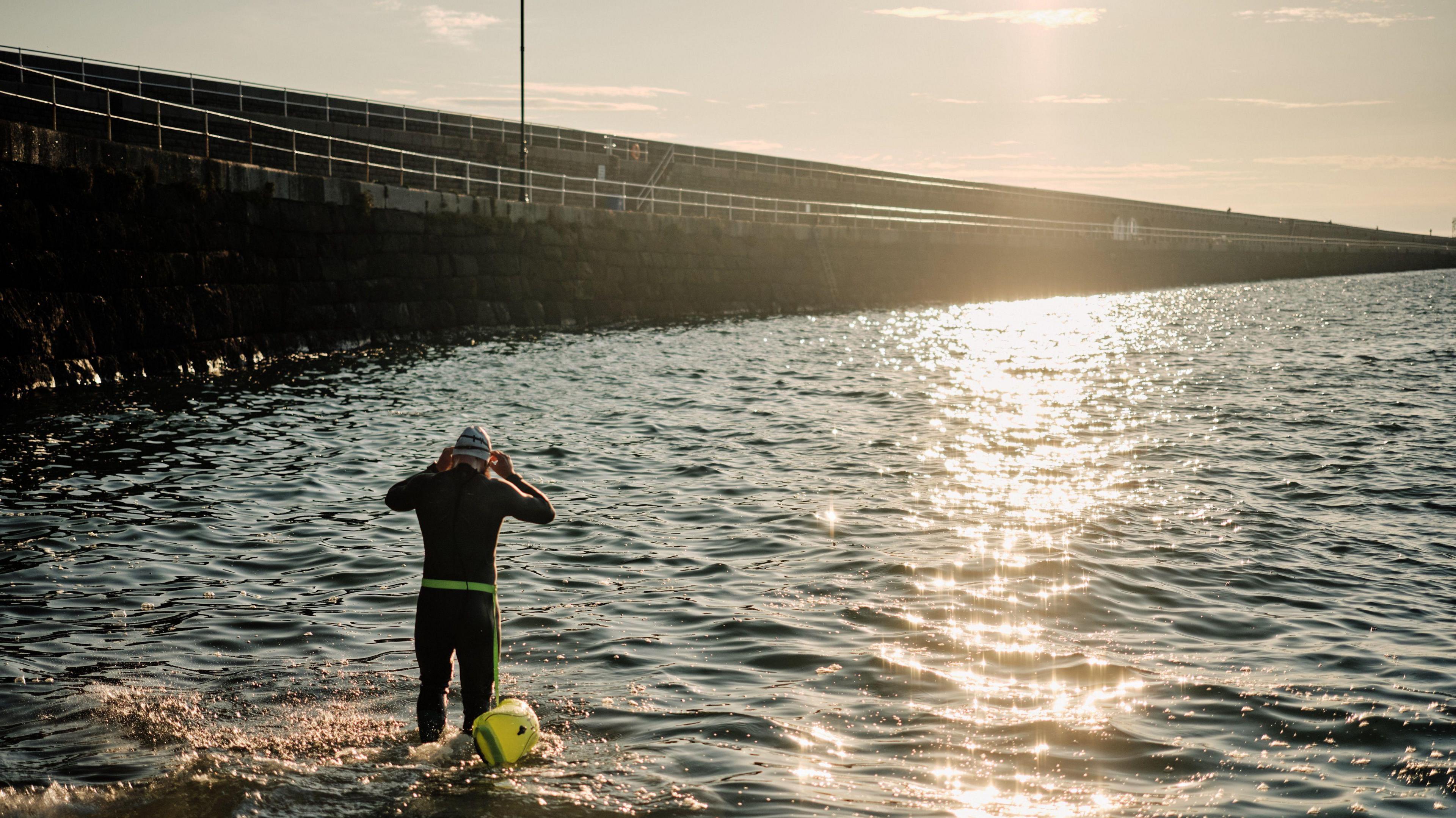 Joe is entering the sea in a swimsuit with his back to the camera as the sun rises over the breakwater. There are slight waves in the sea and Joe has a swim cap on with a safety buoy tied to his waist.