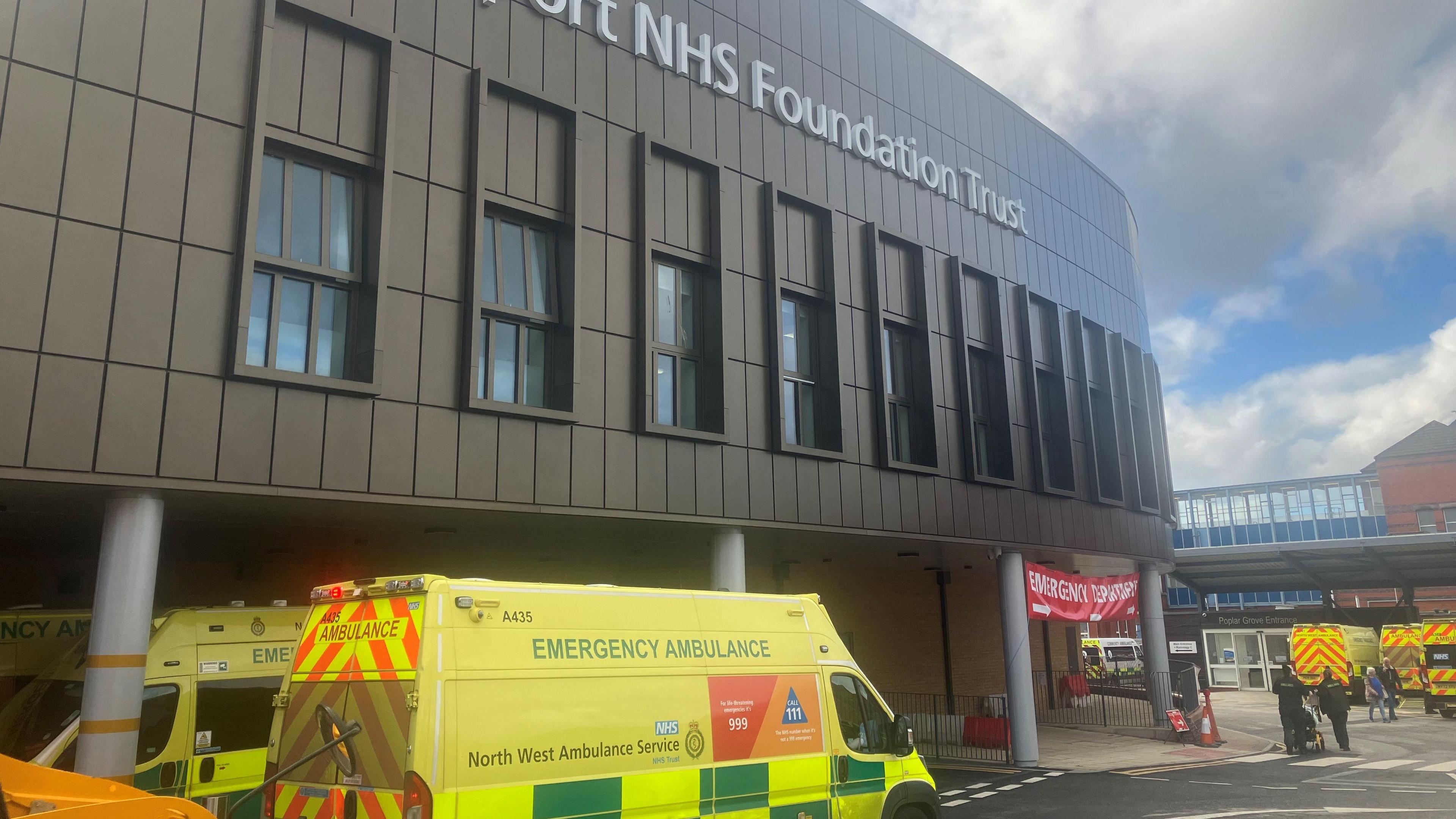 An ambulance at the entrance to Stepping Hill hospital in Stockport, with the main hospital building seen above a roadway and entrance signs and buildings. 