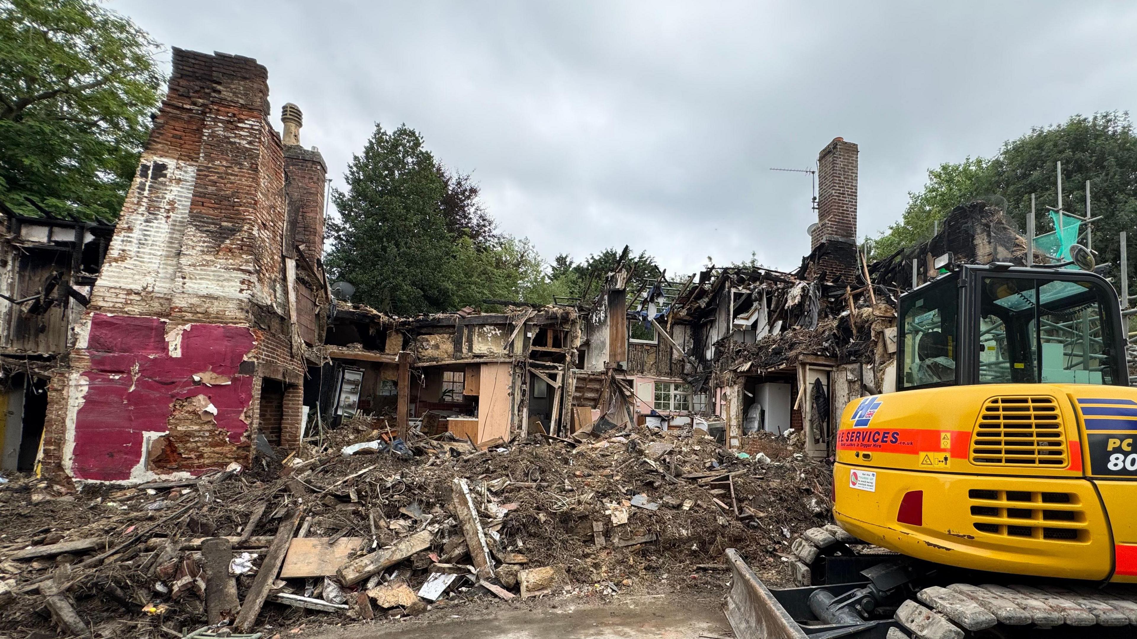A row of cottages completely destroyed with piles of rubble and debris on the floor. You can see inside some of the properties. A digger can be seen to the right of the image. 