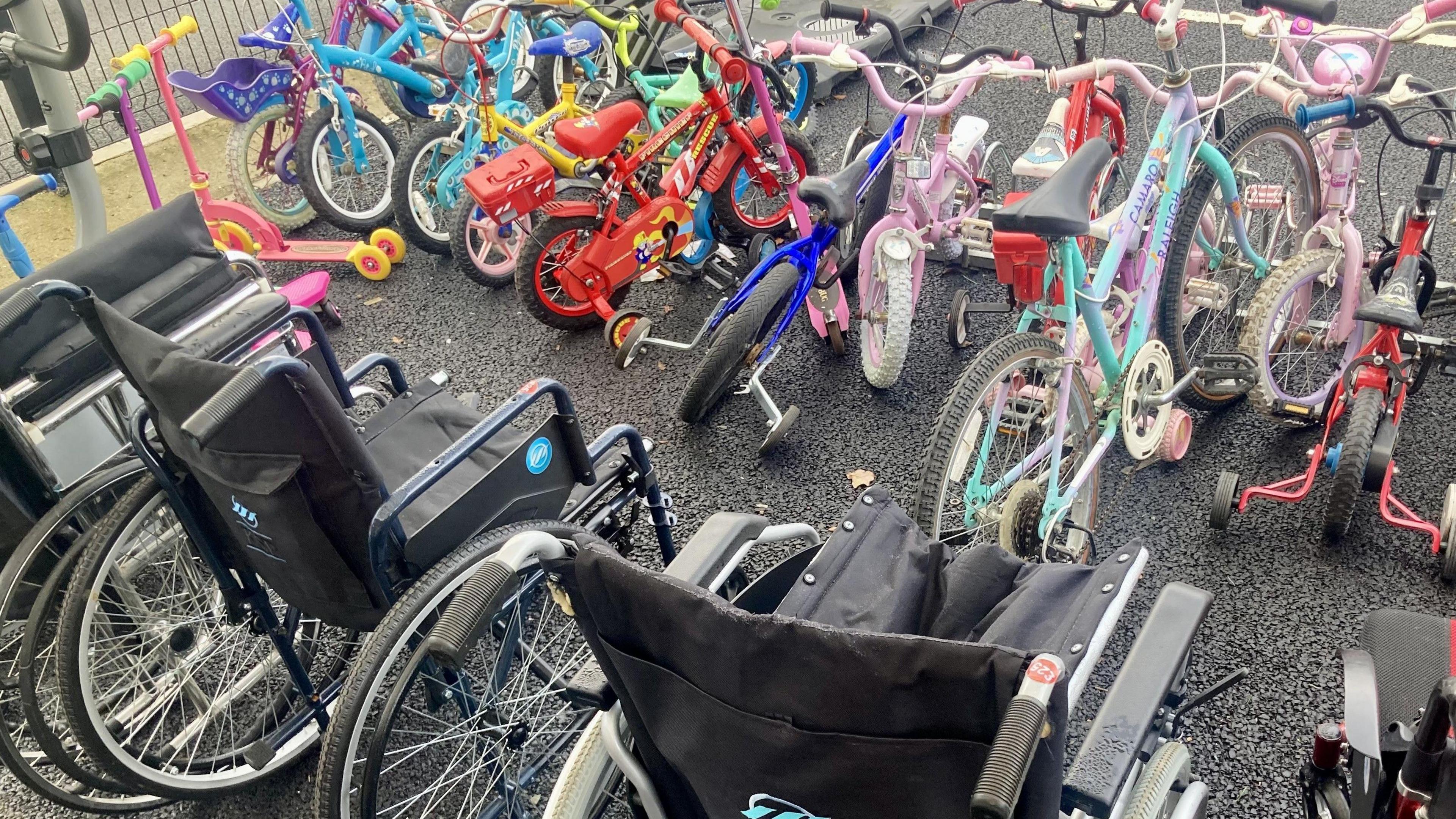 Four black wheelchairs are pictured lined up next to each other behind children's bikes of varying colours.