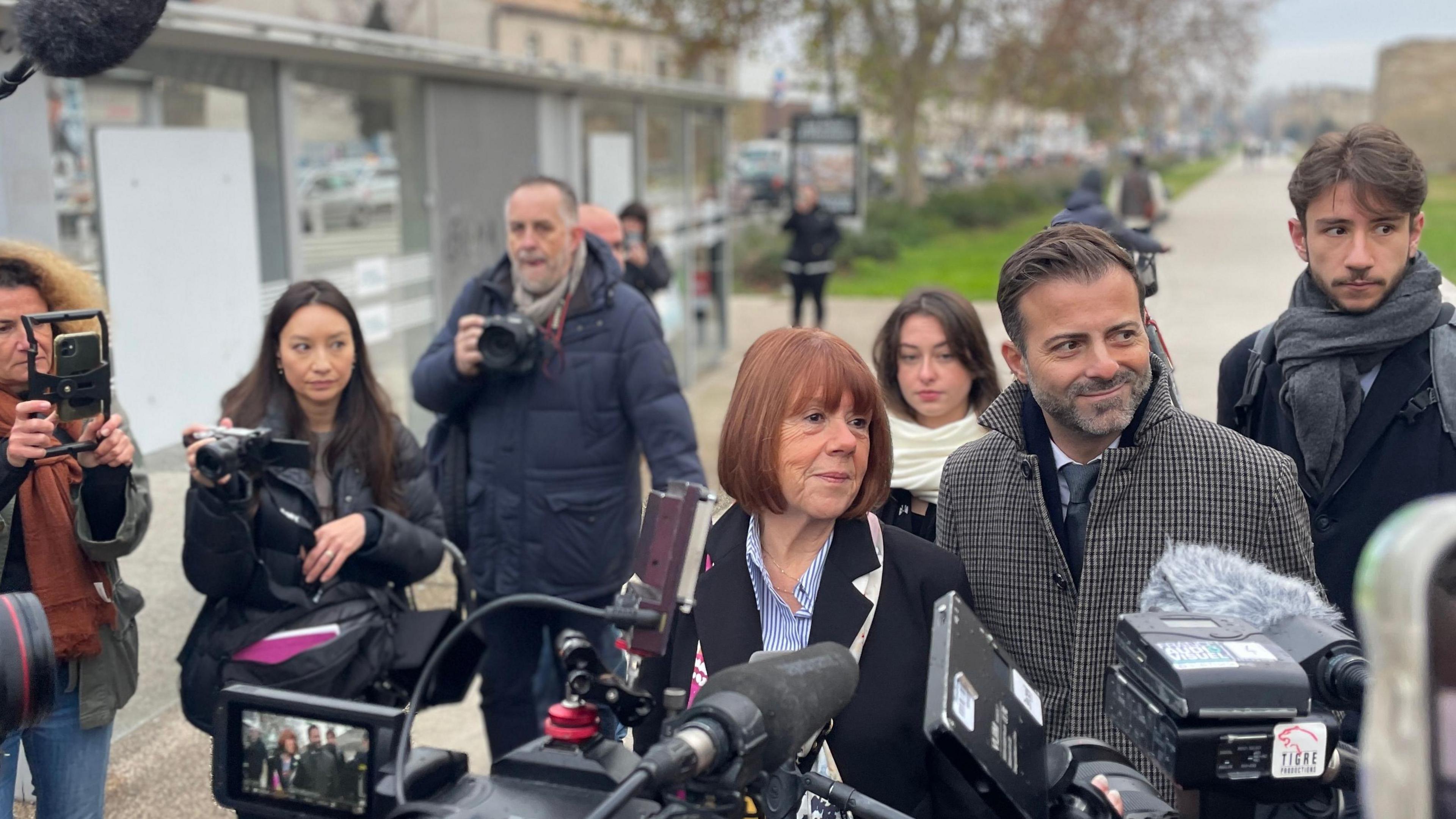 Gisèle Pelicot is surrounding by members of the press carrying cameras. She is walking with her head held high with a faint smile. She has a red bob and is wearing a black coat. She is flanked by her legal team. 