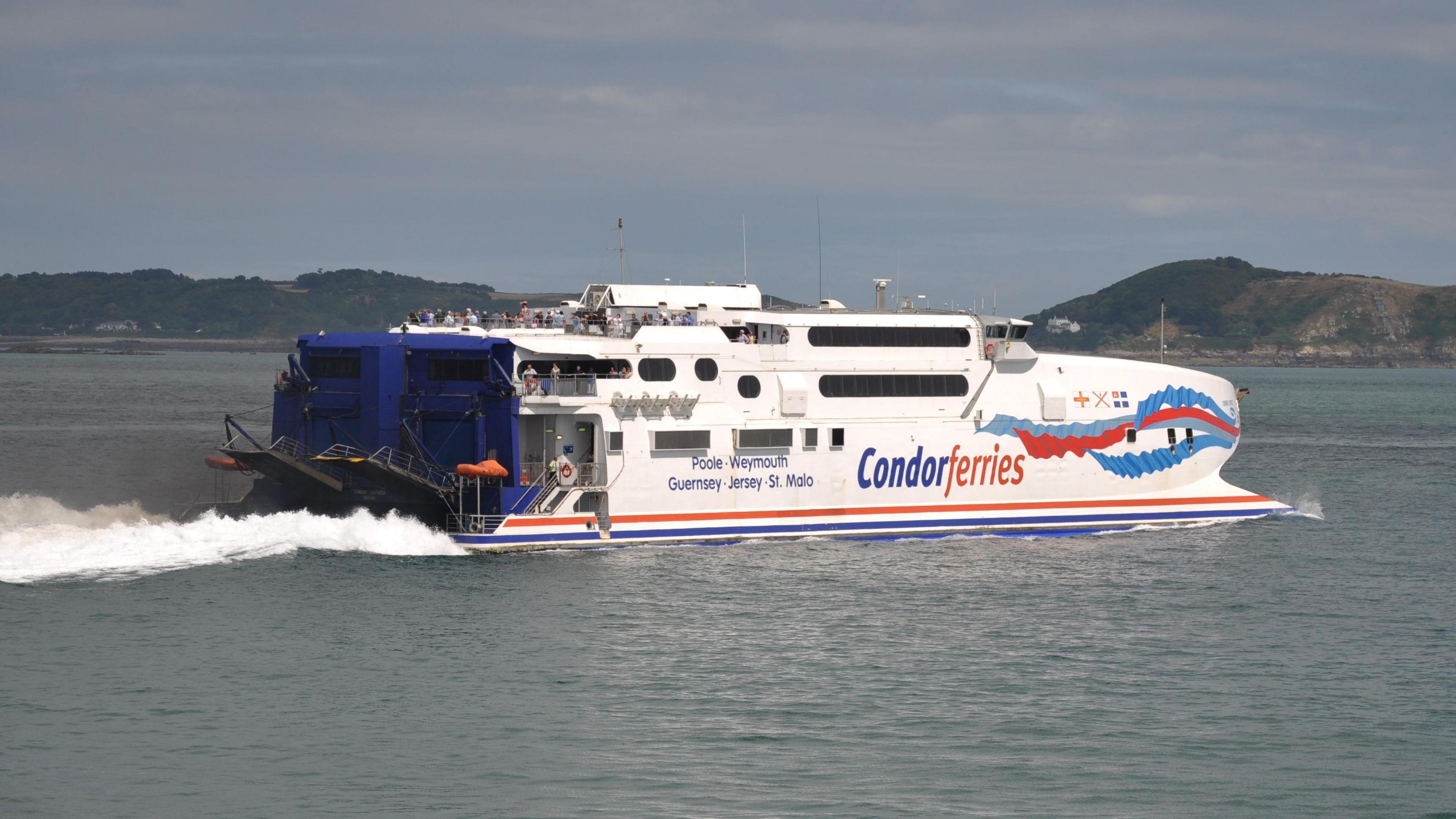 A white, blue and red ferry moving away from the camera with a small wake behind. In the background are two islands - Herm and Jethou