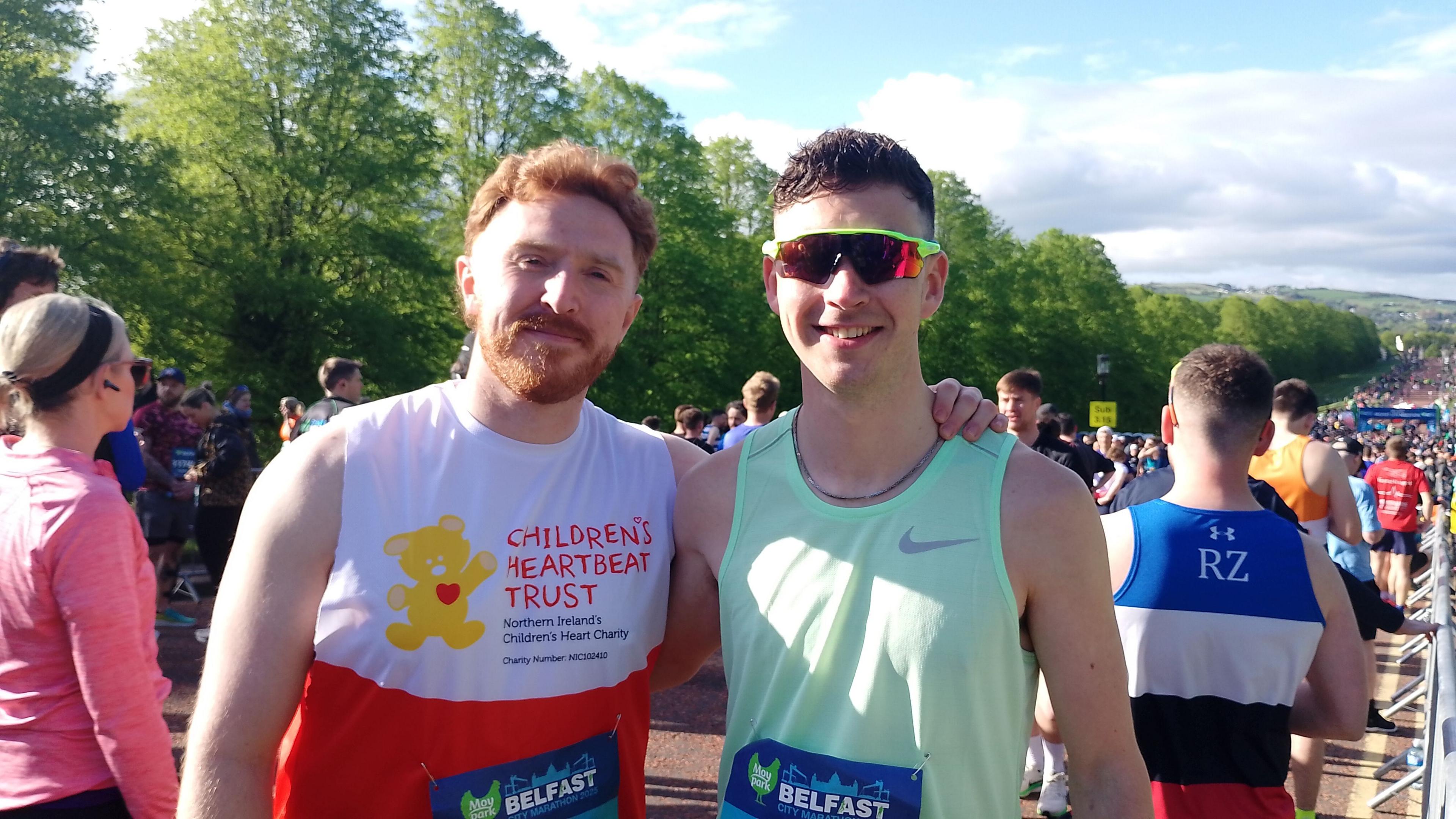 Mr Tolan, who is on the right, stands next to another man. He has short black hair and is wearing a mint green vest with glasses. To the left is a man with ginger hair and facial hair wearing a red and white running vest with 'Children's Heartbeat Trust' written on it.