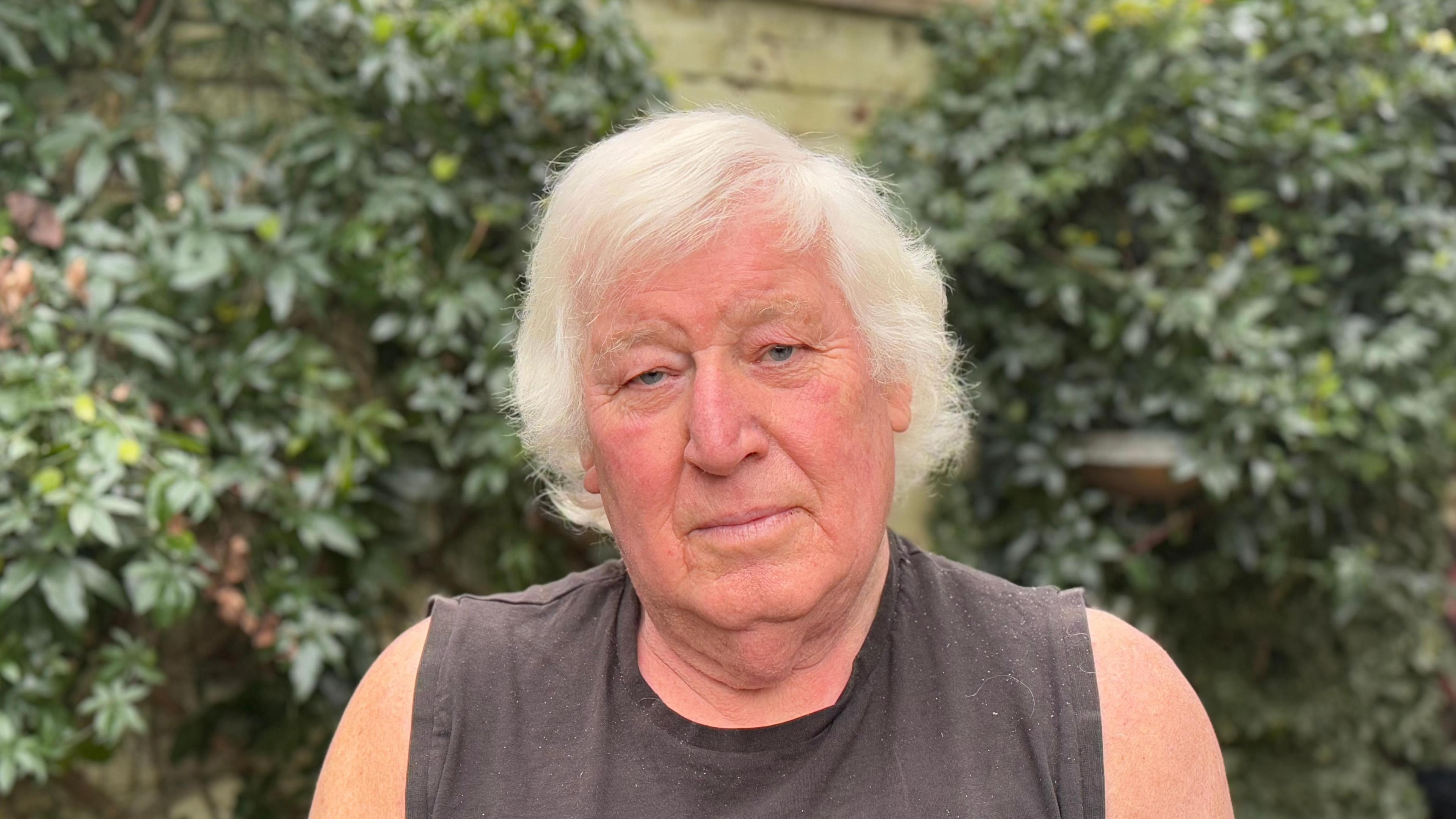 An older man with white hair wearing a vest and standing in front of a garden wall with green leaves on it