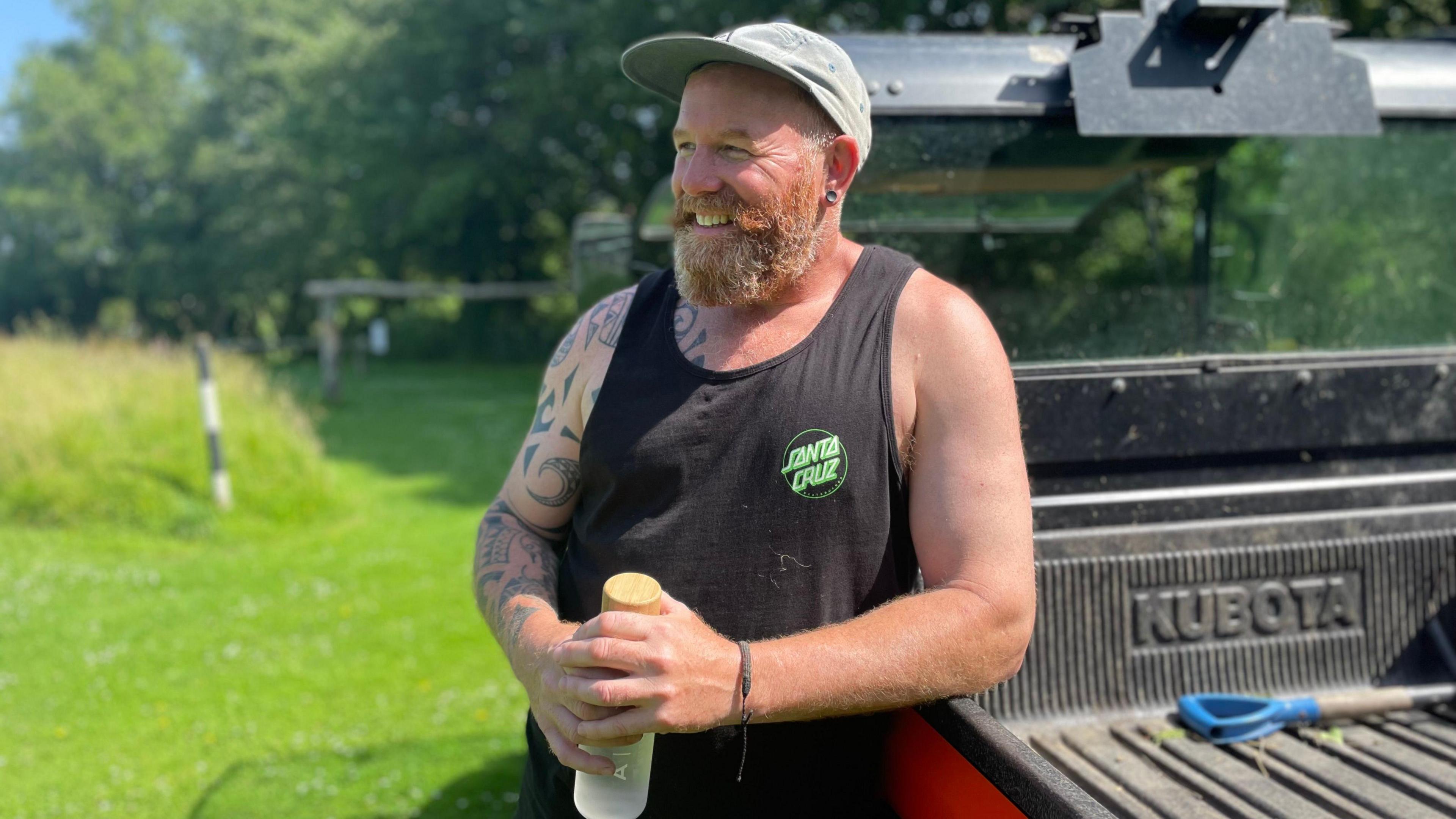 A bearded muscly man wears a sleeveless singlet and a baseball cap, leaning on a small tractor