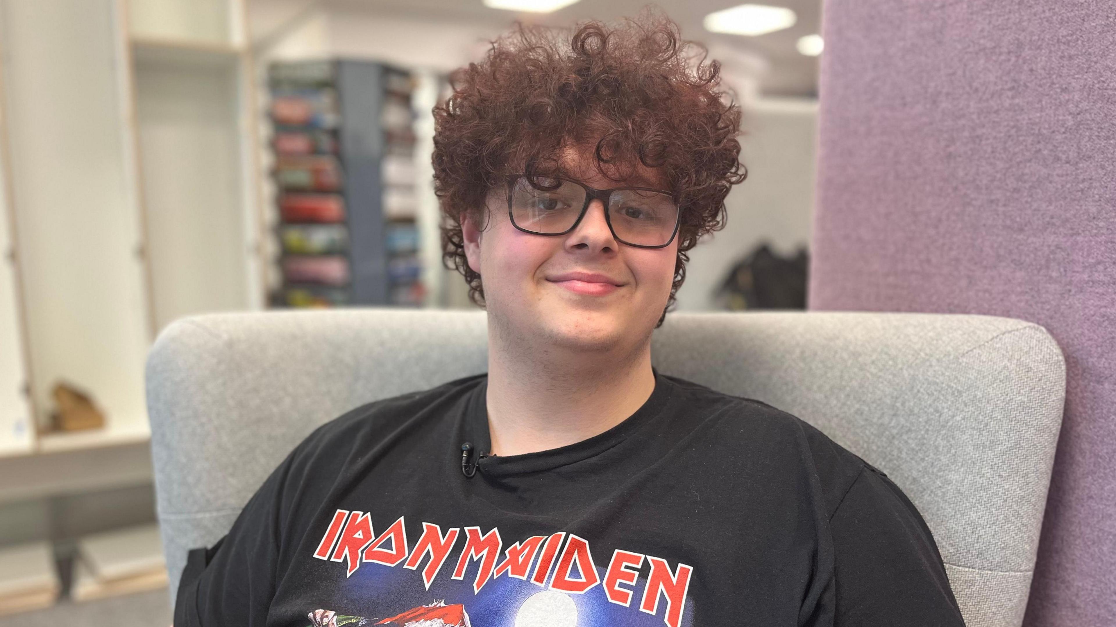 Cameron sydd yn gwisgo crys T du Iron Maiden. Mae ganddo wallt cyrliog a sbectol