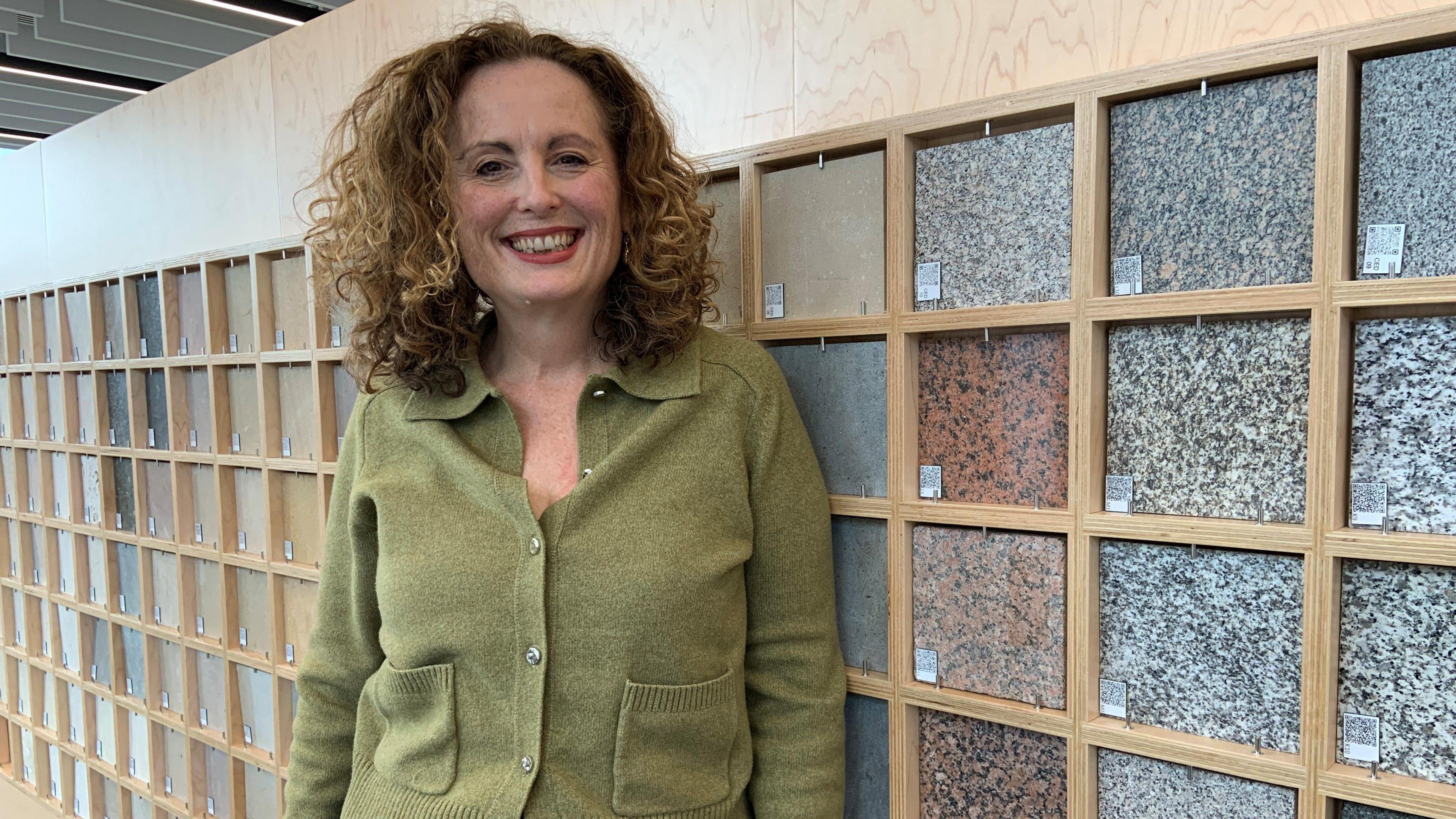 A woman with brown, shoulder length curls is smiling. She is wearing a moss green cardigan and is stood in front of a wall with dozens of different coloured stone tiles