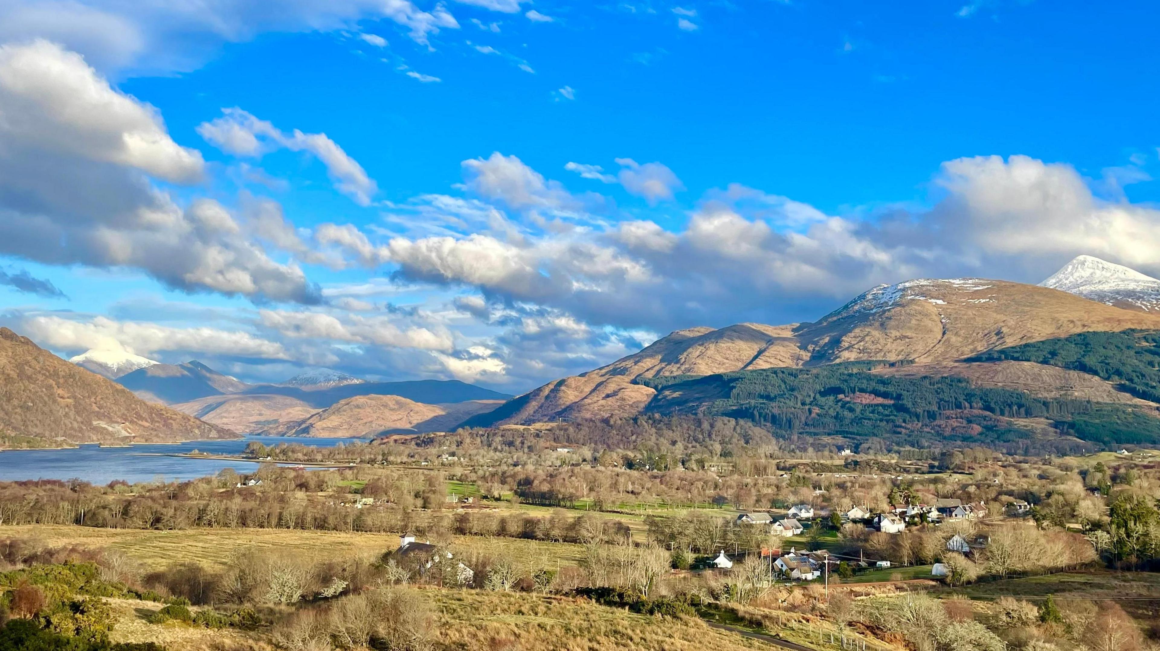 Landscape picture of the Scottish Highlands with fields and a village of white painted houses in the foreground.  Mountain peaks with snow on top are set against a background of a sunny blue sky filled with wispy white clouds.