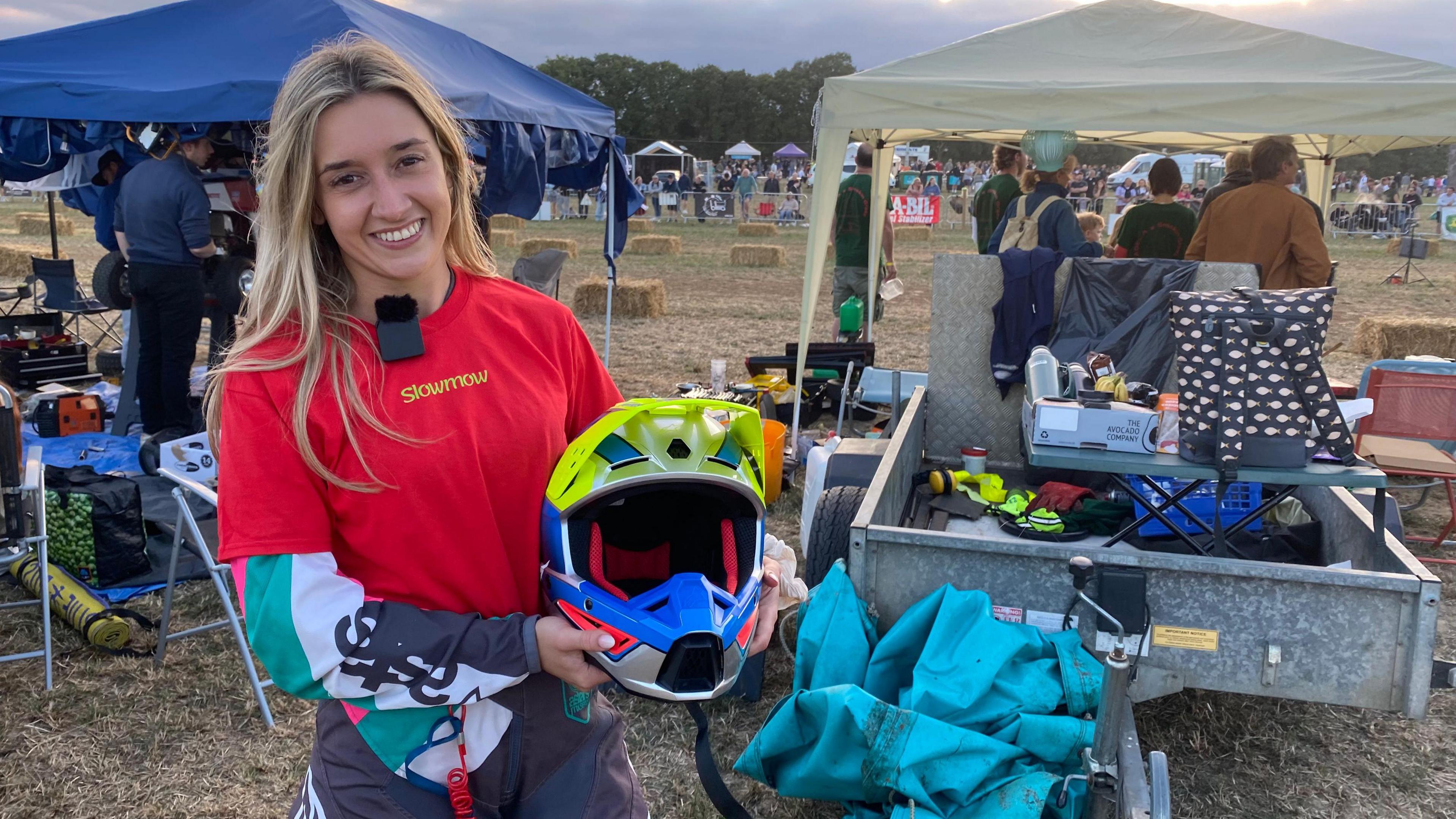 A woman, in her racing uniform and holding a helmet, is smiling at the camera. A dirt race track is behind her