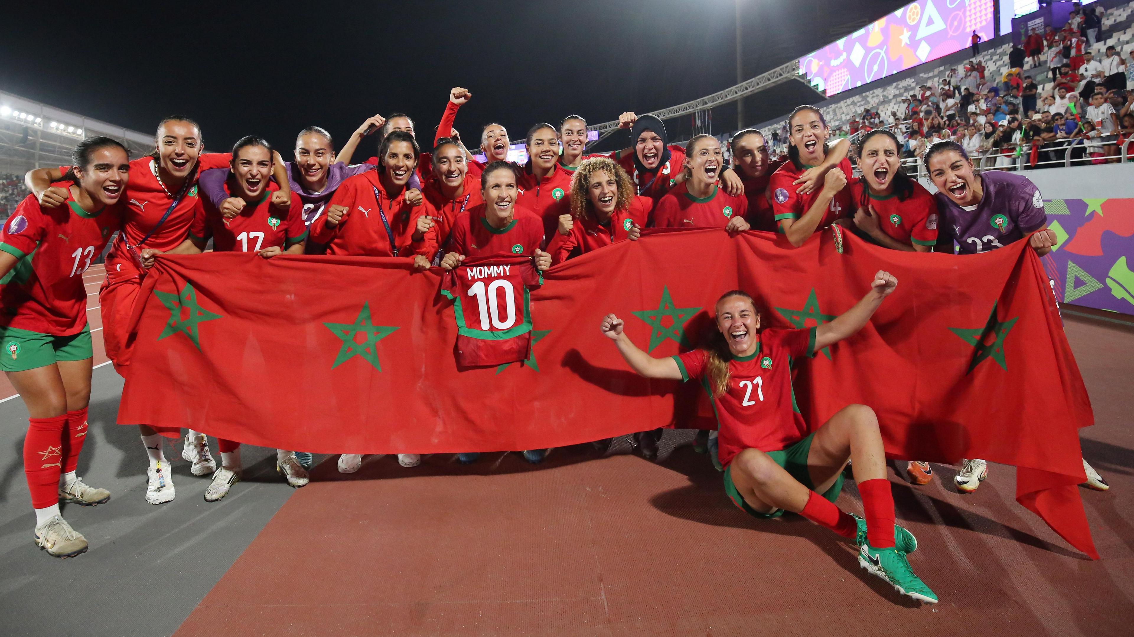 Morocco players celebrate inside a stadium after winning their Women's Africa Cup of Nations semi-final by gathering around a large red banner made up of several Moroccan flags joined together, with most players standing behind it, while one of them hangs a mini kit over the top that shows the number 10 and the word mommy on its back