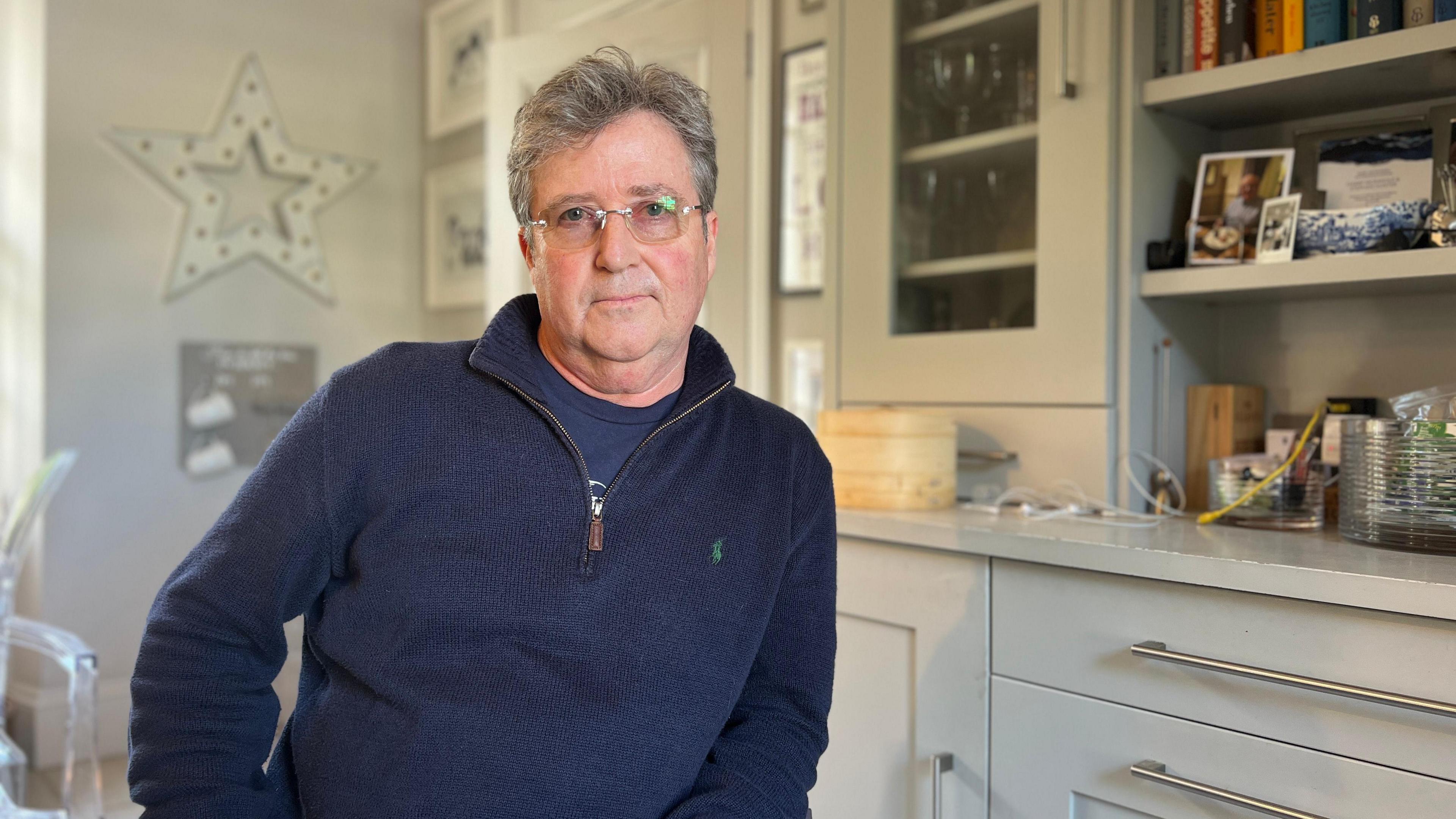 Jason looks at the camera as he sits in a kitchen. Jason is wearing a blue, knitted zip up jumper with a dark blue t-shirt under it. He is wearing glasses with square frames. There's a cabinet to his left with glasses on one side and books on the other side.