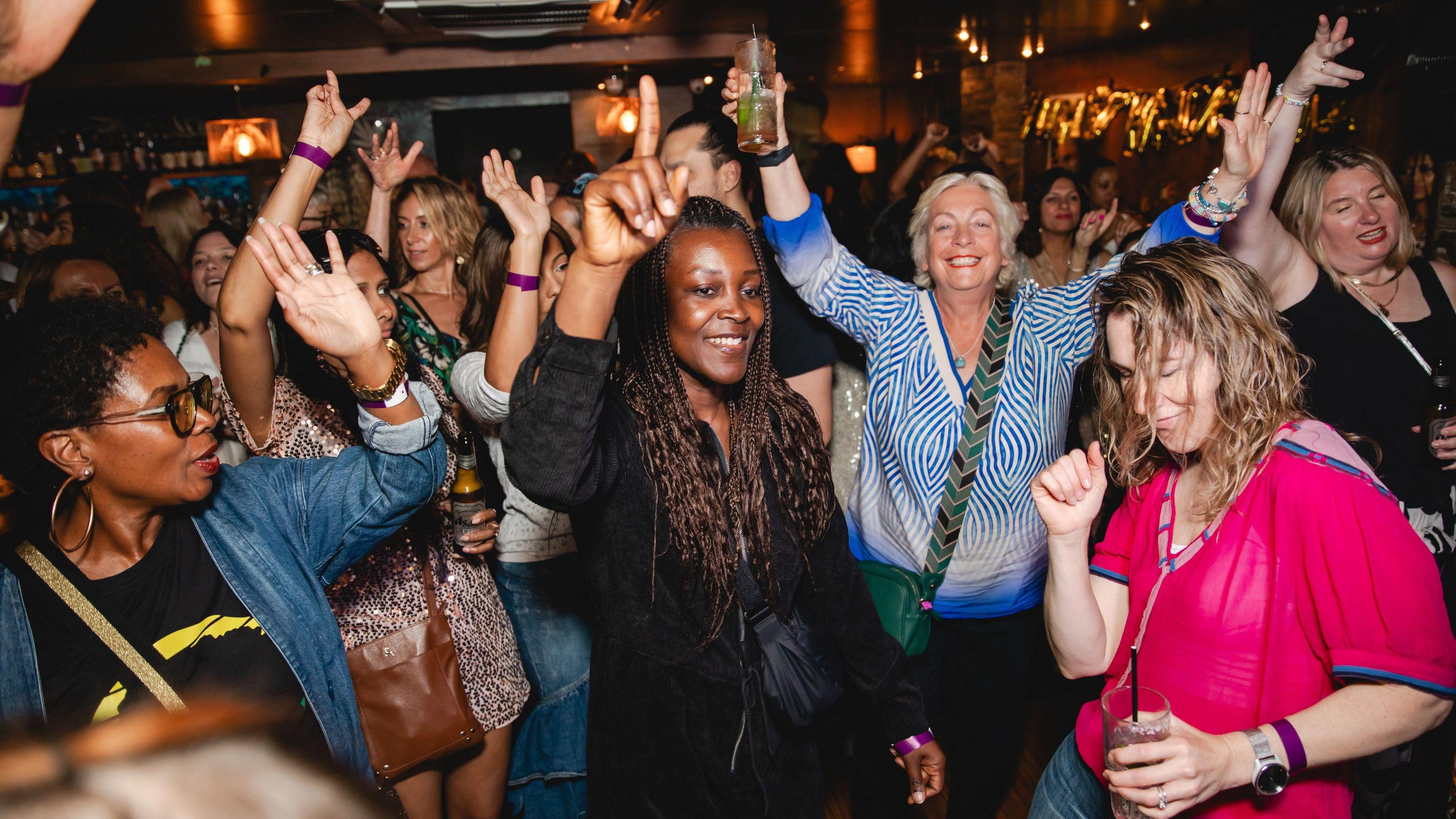 A group of women of a range of ages dancing in a room. Some of them have their hands in the air and are holding drinks.