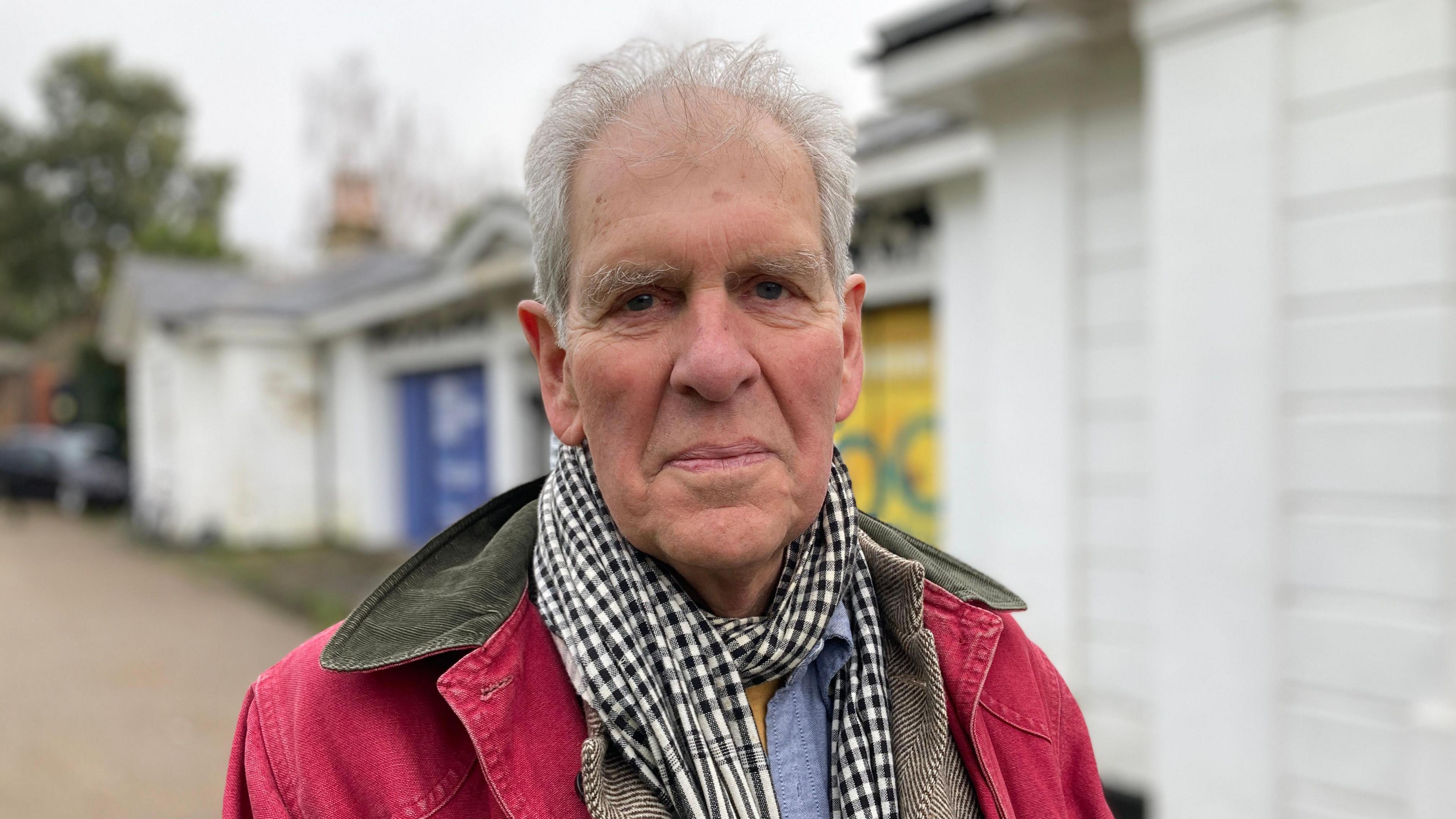 Alastair Sawday wears a black and white checked scarf and a red jacket. He has short white hair and stands outside of Bristol Zoo Gardens.