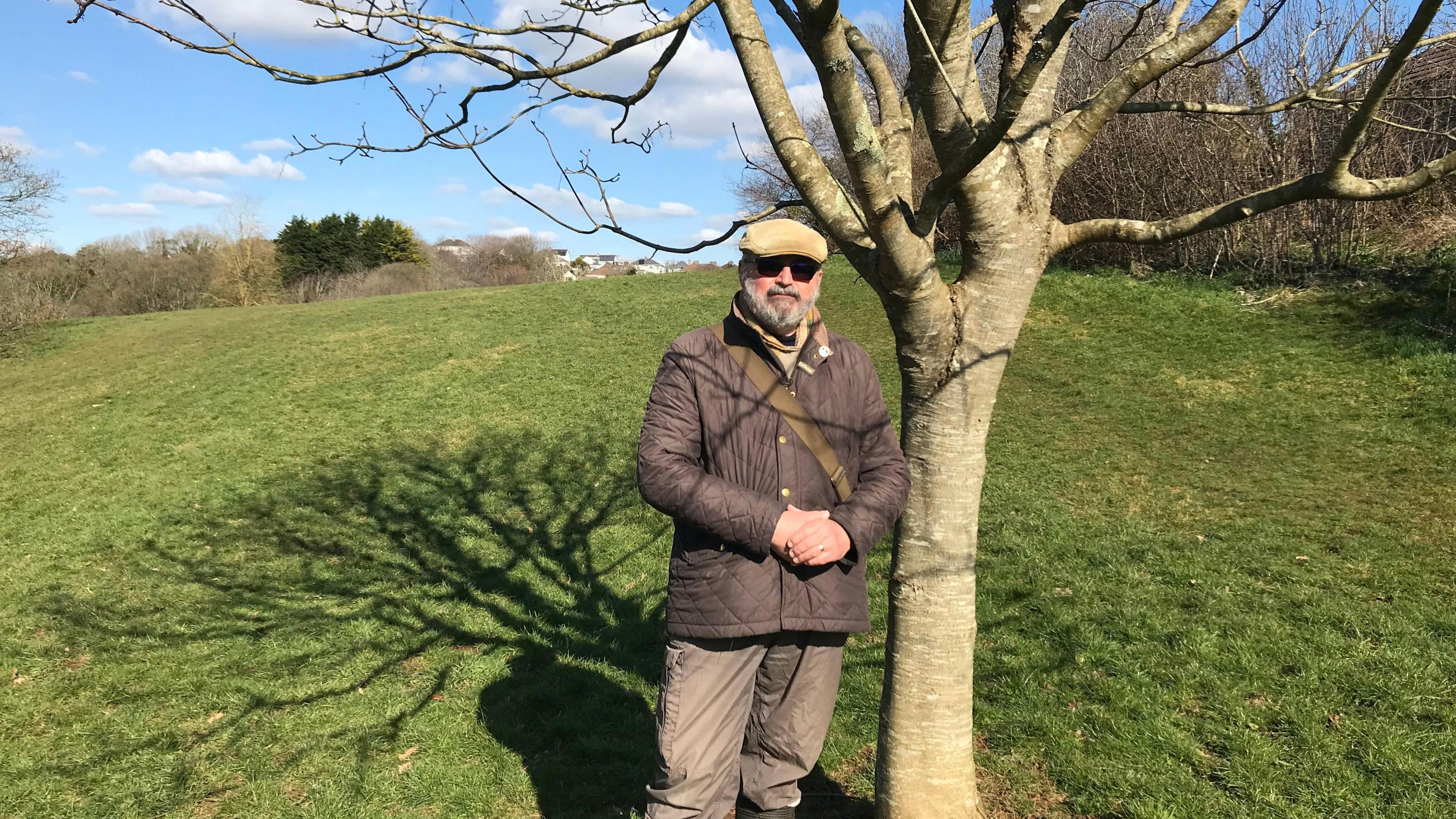 Kevin Warley, a man with a grey beard in sunglasses standing beside his father's memorial tree.