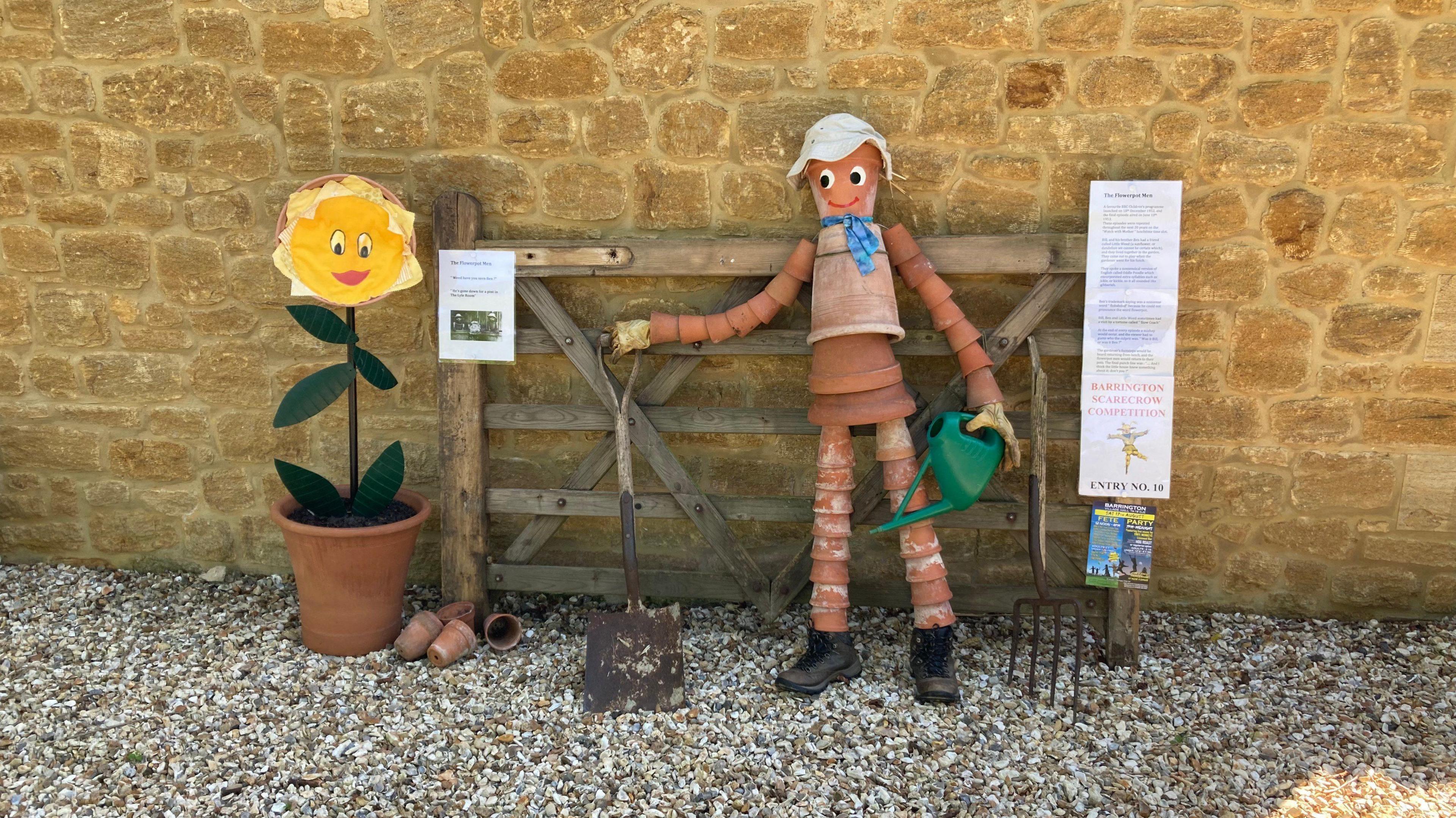 A scarecrow made out of plant pots. Googly eyes and a smile have been put on it, and a flower scarecrow with a face is next to it. 