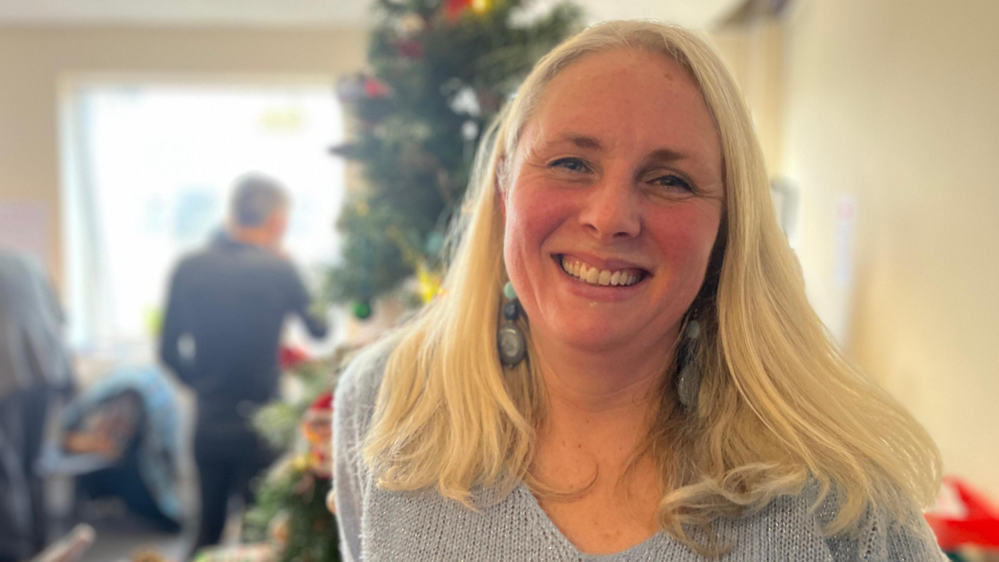 Nikki Packham stands in front of a Christmas tree. She has blonde hair and is smiling. She is wearing earrings.