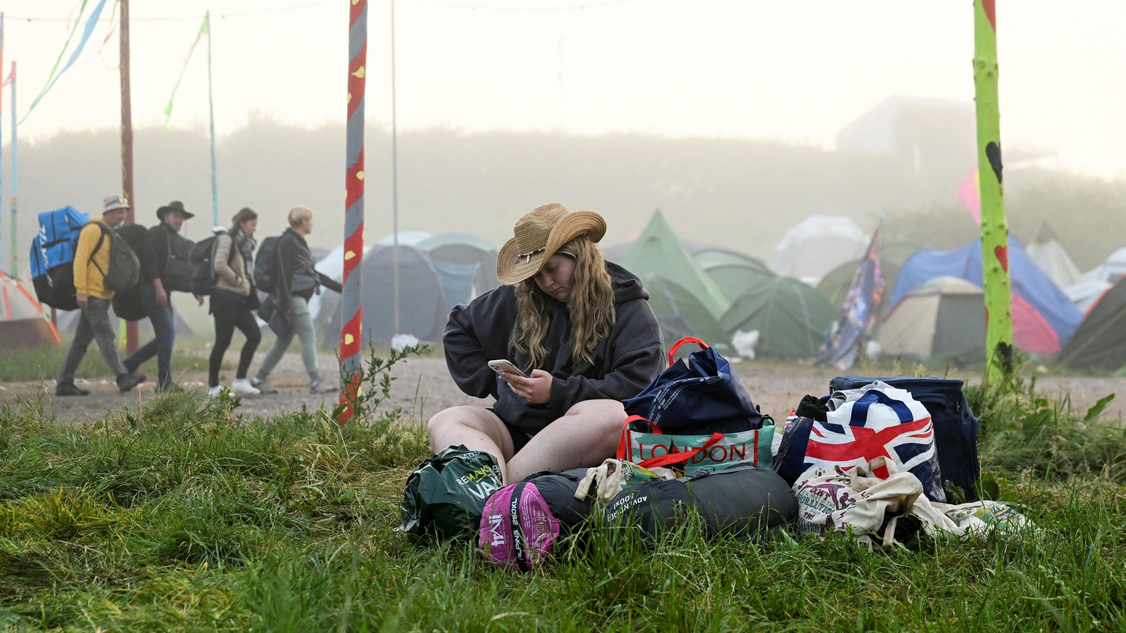 A woman sat on the grass at Glastonbury Festival. She is wearing a black jacket, shorts and a brown cowboy hat. She is looking down at her phone. Her belongings are bagged up next to her. A group of punters carrying camping gear and luggage can be seen on the path behind her. In the distance, there are many tents.